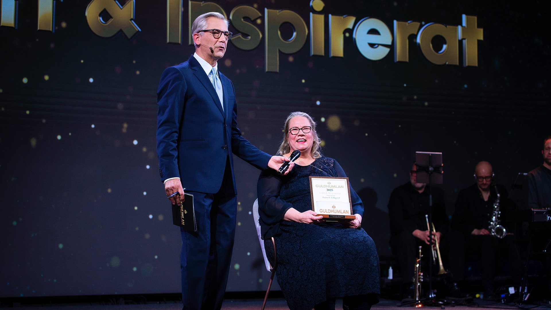 En man i mörk kostym och en sittande kvinna i mörk klädsel vid en prisutdelningscermoni. Kvinnan håller ett diplom eller certifikat. I bakgrunden syns en scen med musikanter och en stor skärm.