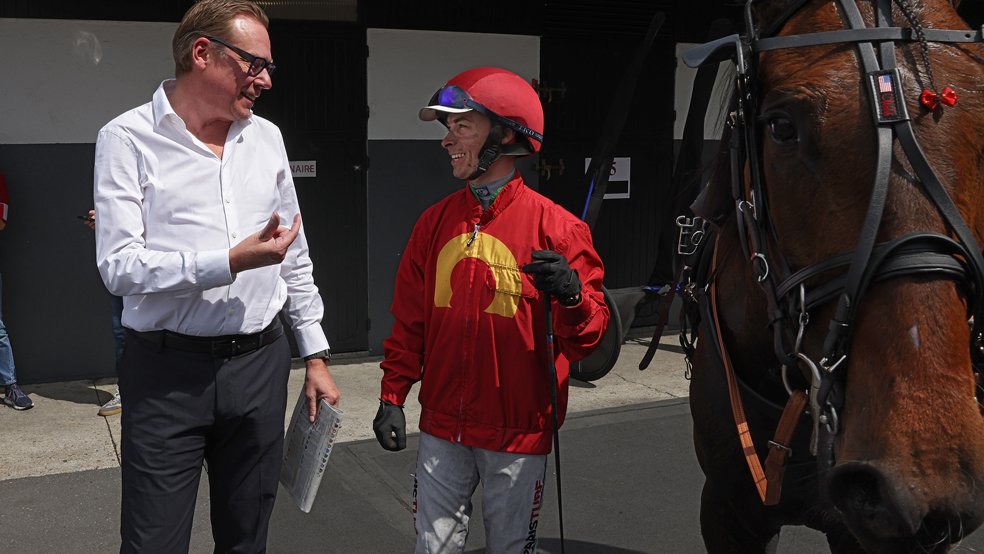 En man i vit skjorta samtalar med en jockey i röd jacka och hjälm vid en brun häst.