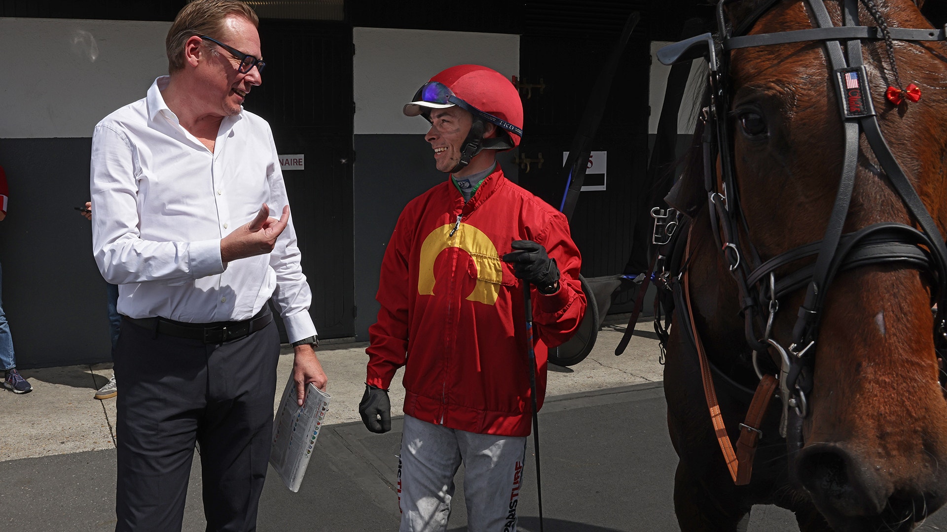 En man i vit skjorta samtalar med en jockey i röd jacka och hjälm vid en brun häst.