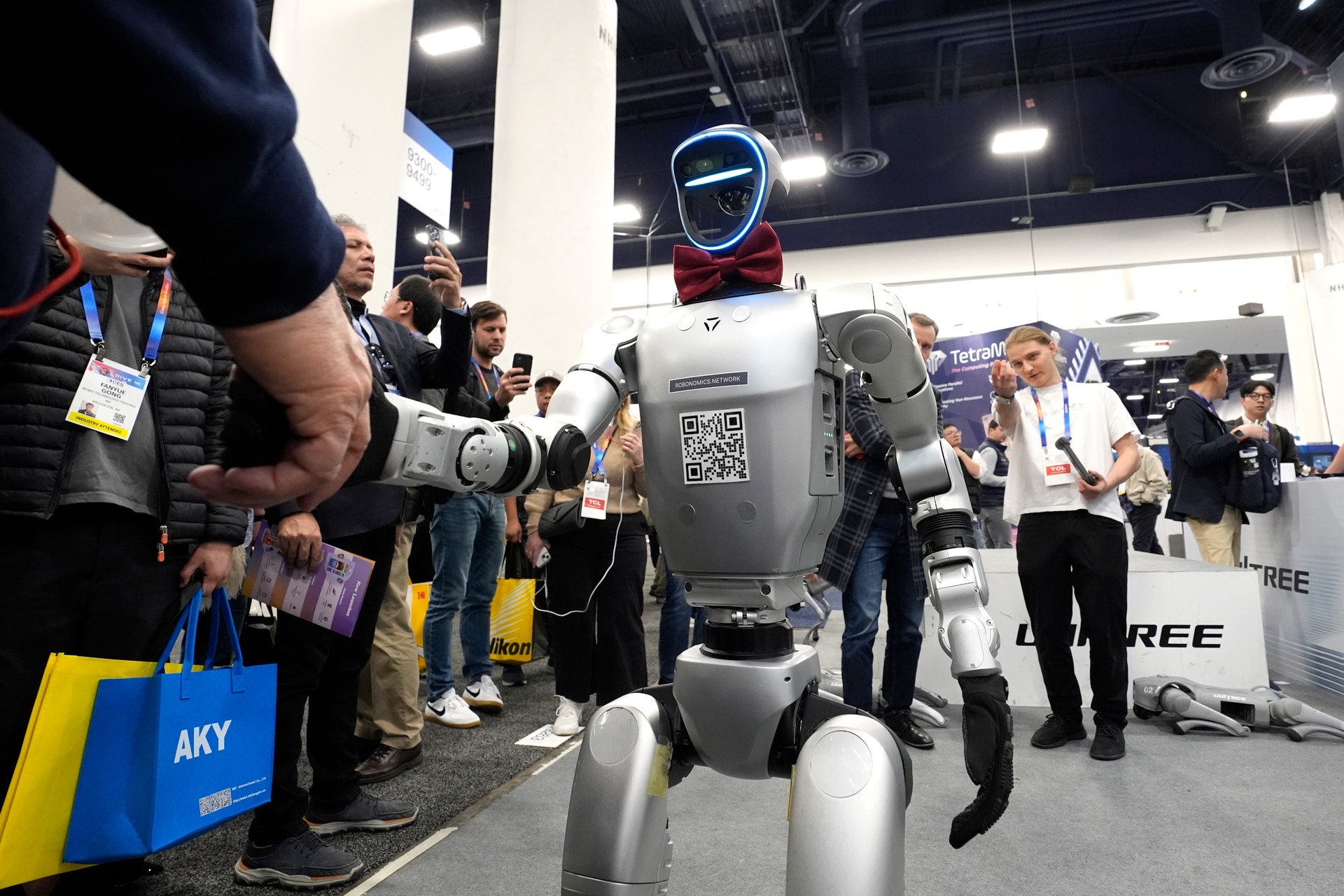 En humanoid robot med vit och grå kropp och blått huvud står på en mässa omgiven av människor som fotograferar och observerar.