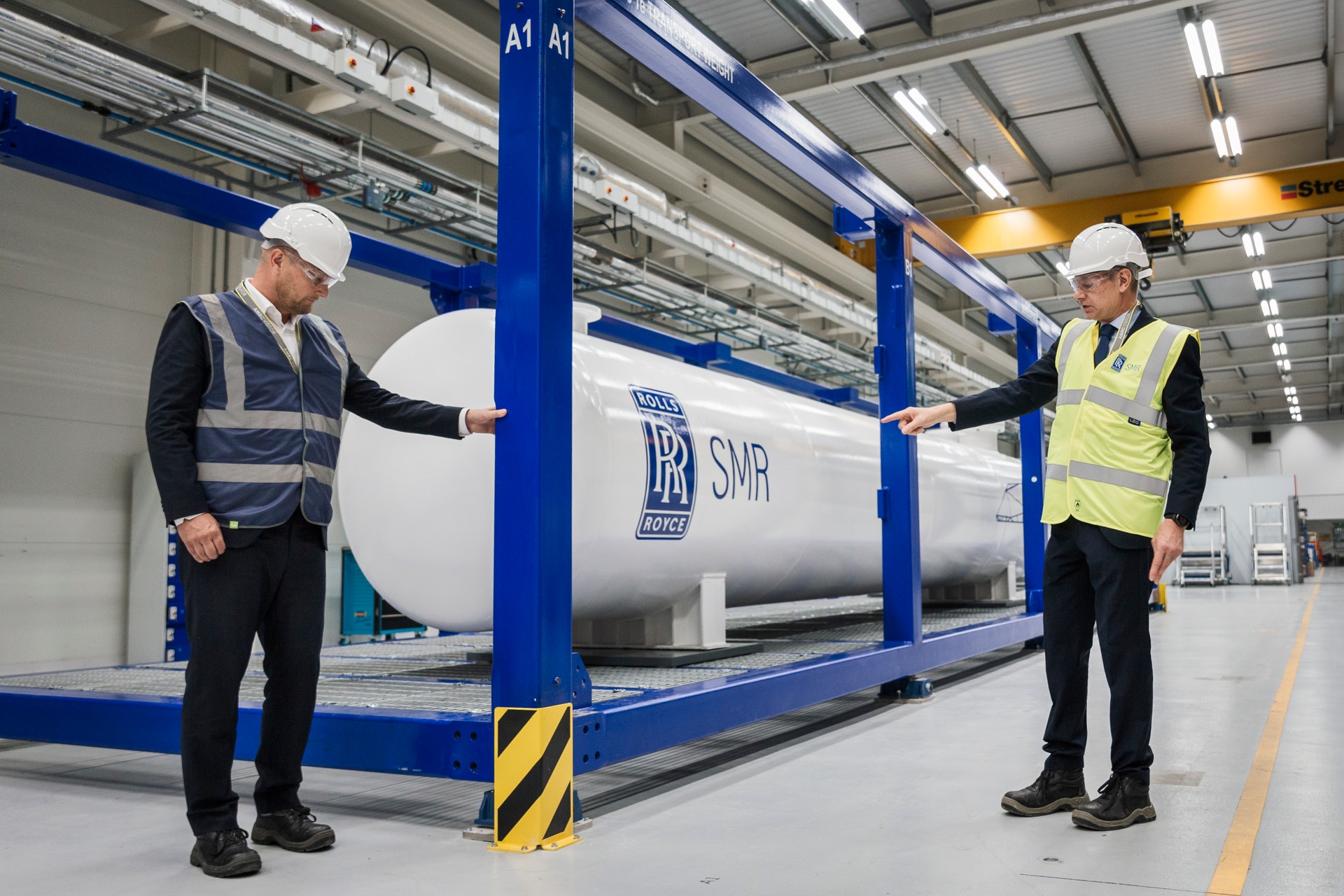 Två arbetare i säkerhetsutrustning står vid en stor cylindrisk industriell tank i ett fabrikslager.