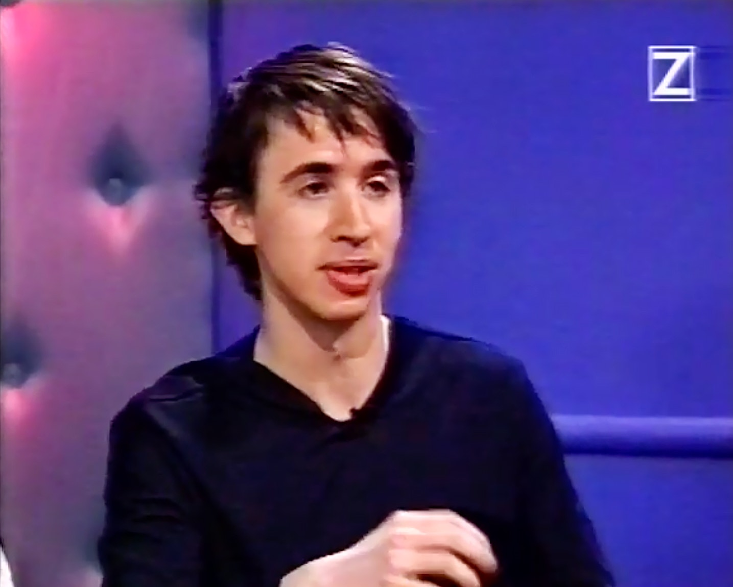 En person i en svart tröja sitter och talar på en tv-studio med blå och rosa bakgrund.