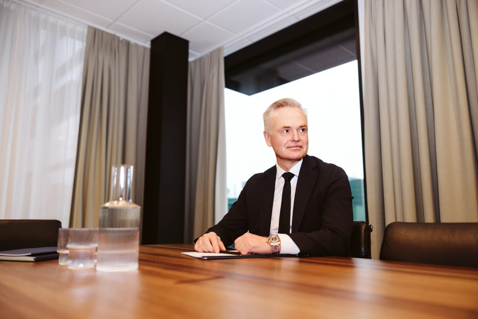 Man i svart kostym och slips sitter vid ett träbord i ett modernt kontorsrum med stora fönster och beiga gardiner.