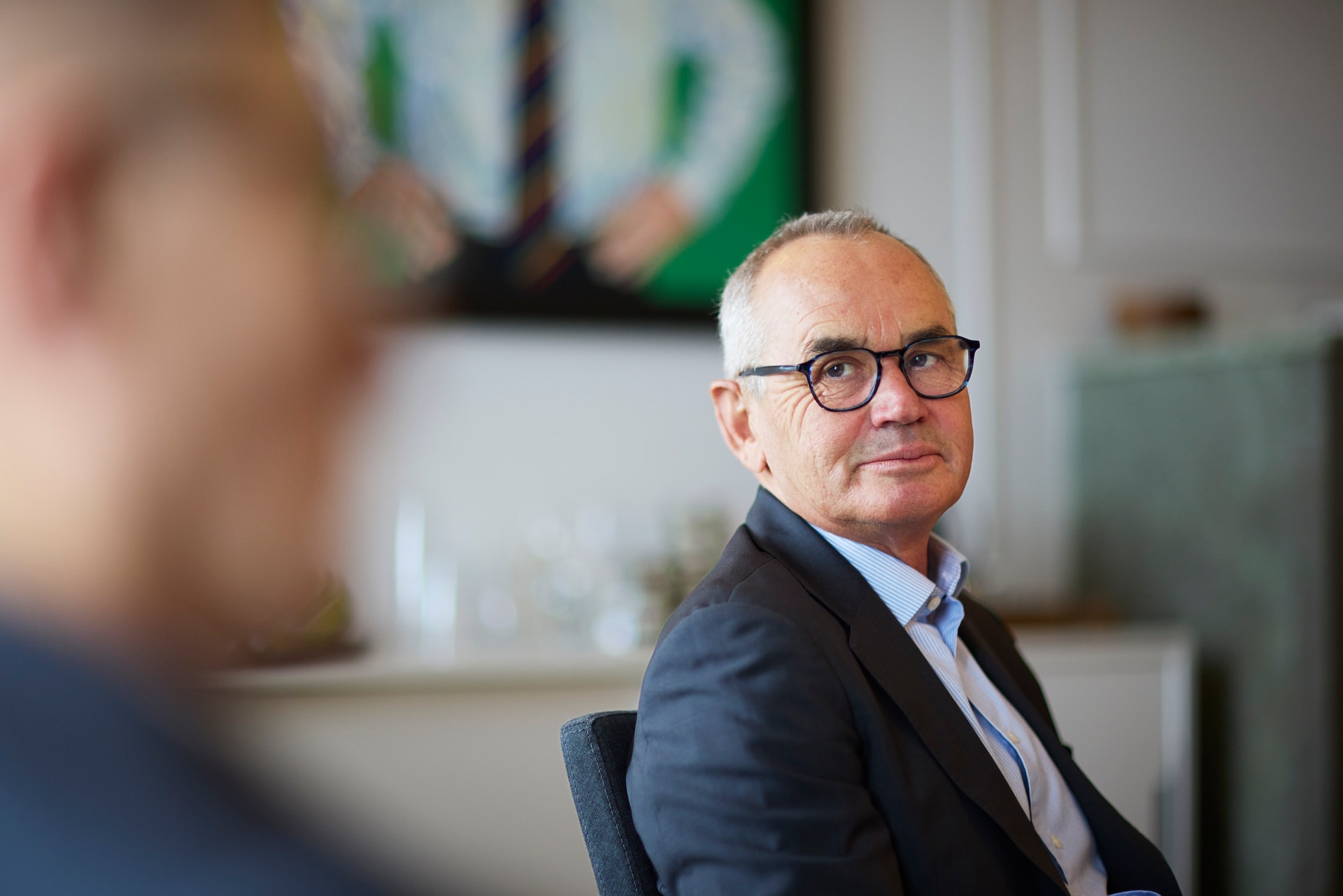 Porträtt av en man med glasögon och vit hår, klädd i mörkblå kavaj, sittande i ett kontor.