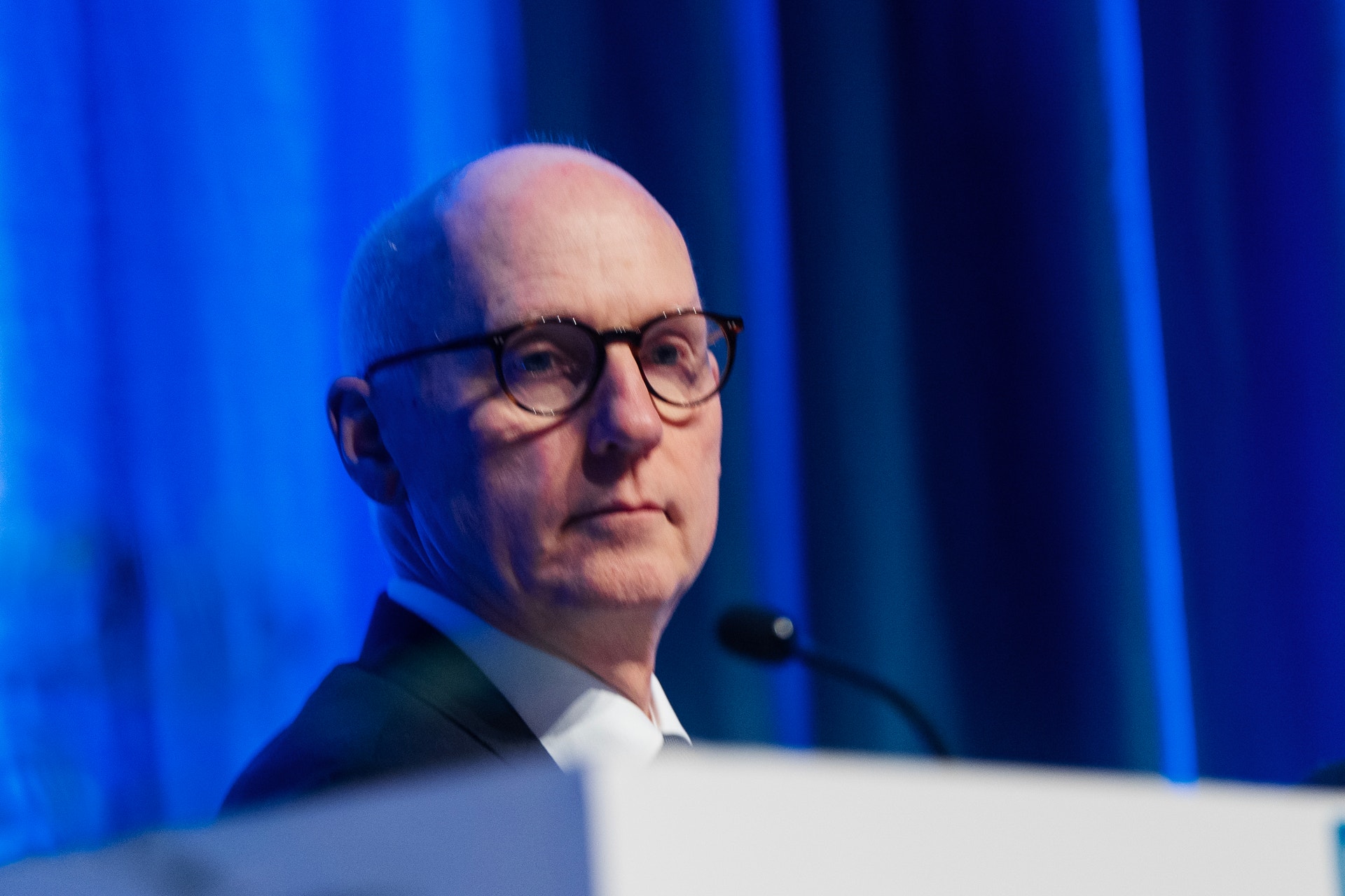 En man med glasögon talar vid ett podium mot en blå bakgrund.