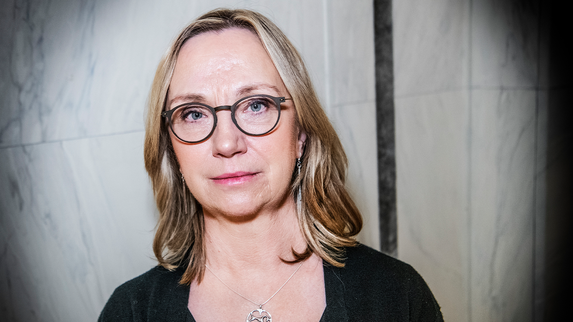 Porträttfoto av en kvinna med glasögon och långt ljusbrunt hår mot en neutral bakgrund.