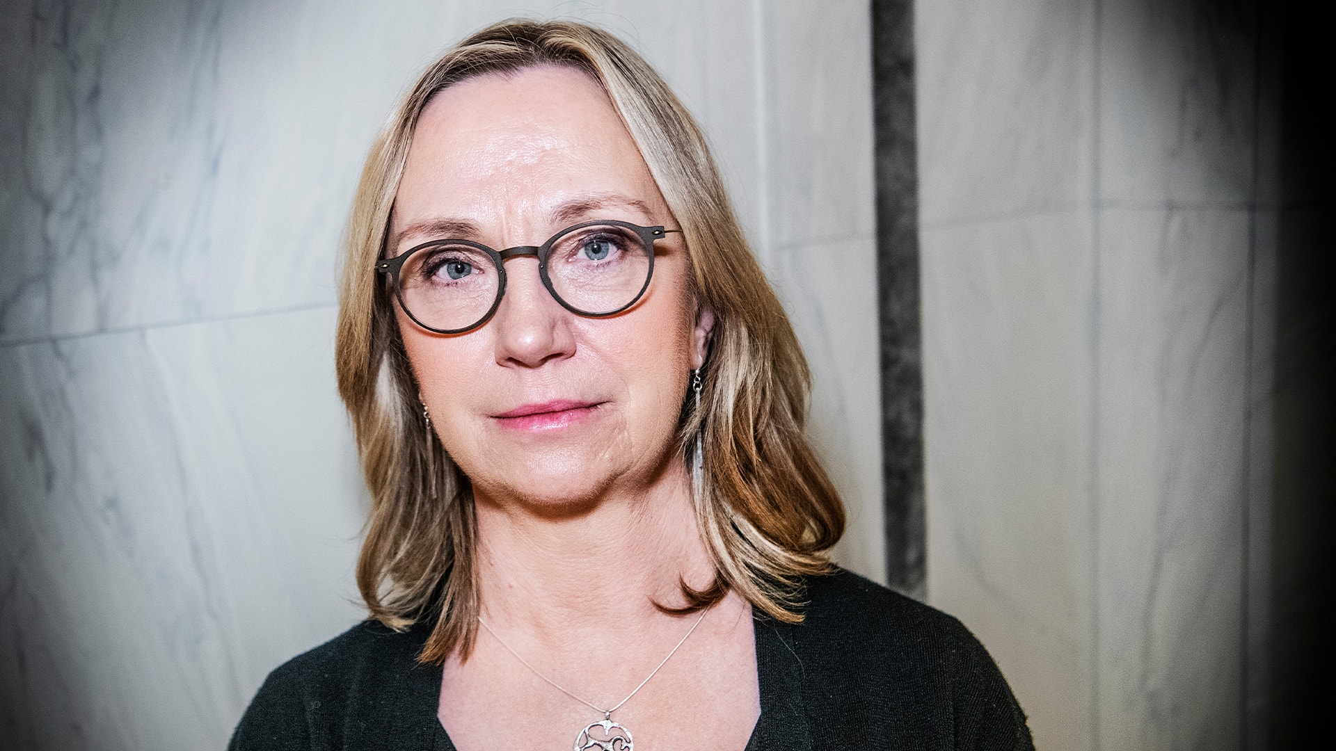 Porträttfoto av en kvinna med glasögon och långt ljusbrunt hår mot en neutral bakgrund.