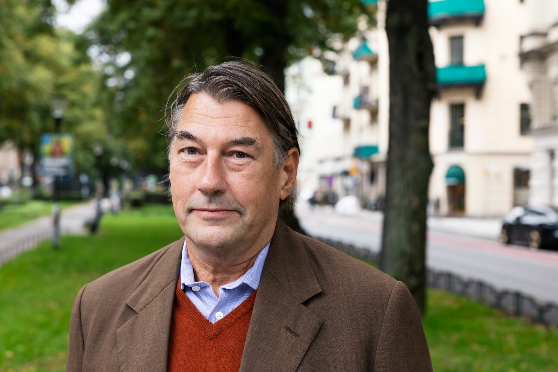 Porträtt av en man i kavaj som står utomhus på en stadsgata med träd och byggnader i bakgrunden.