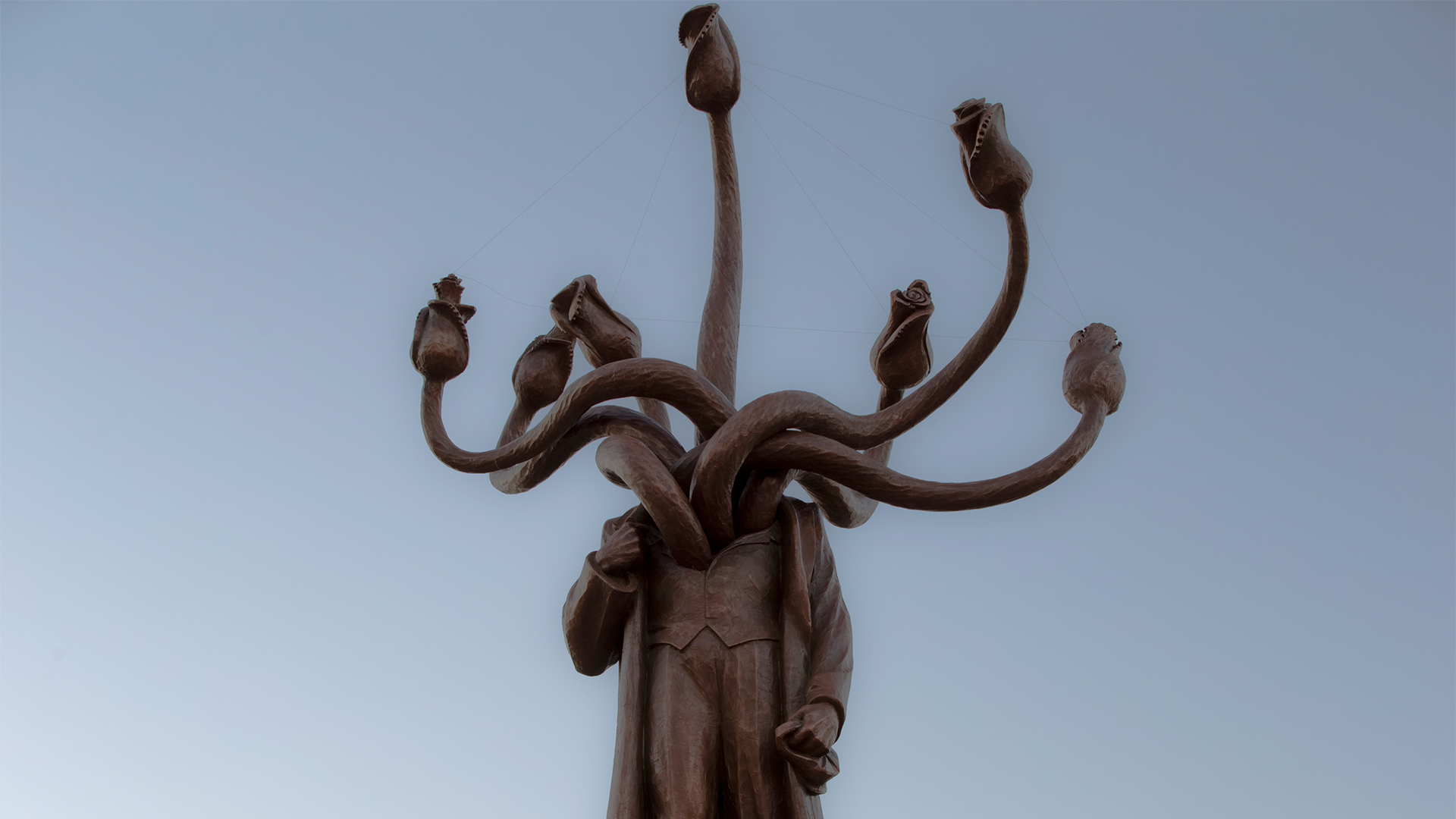 Skulptur av hydran med flera slanglika huvuden som sträcker sig uppåt från en central kropp mot en blå himmel.