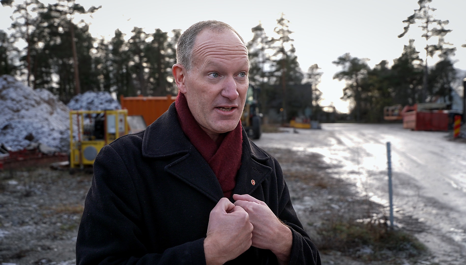 Porträtt av en man som står utomhus i vintermiljö. Han bär svart jacka och röd tröja och verkar prata eller förklara något.