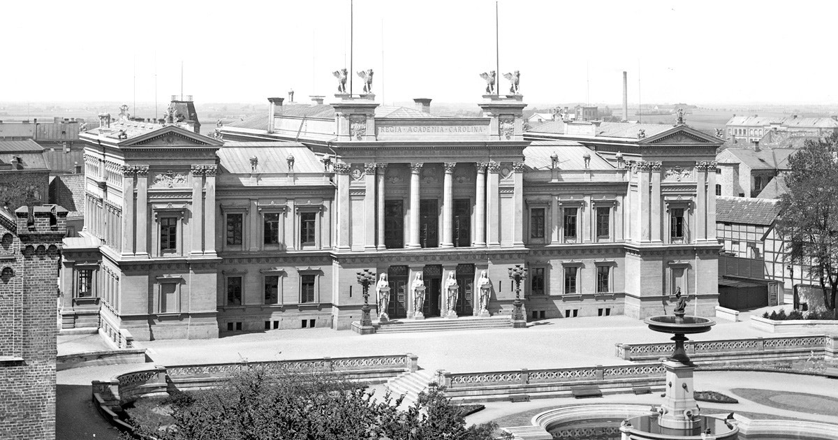 Svartvit historisk fotografi av Lunds universitetsbiblioteks huvudbyggnad från innan 1897. En klassicistisk byggnad med vita kolonner och symmetrisk arkitektur, fotograferad från ovan med omgivande stad i bakgrunden.