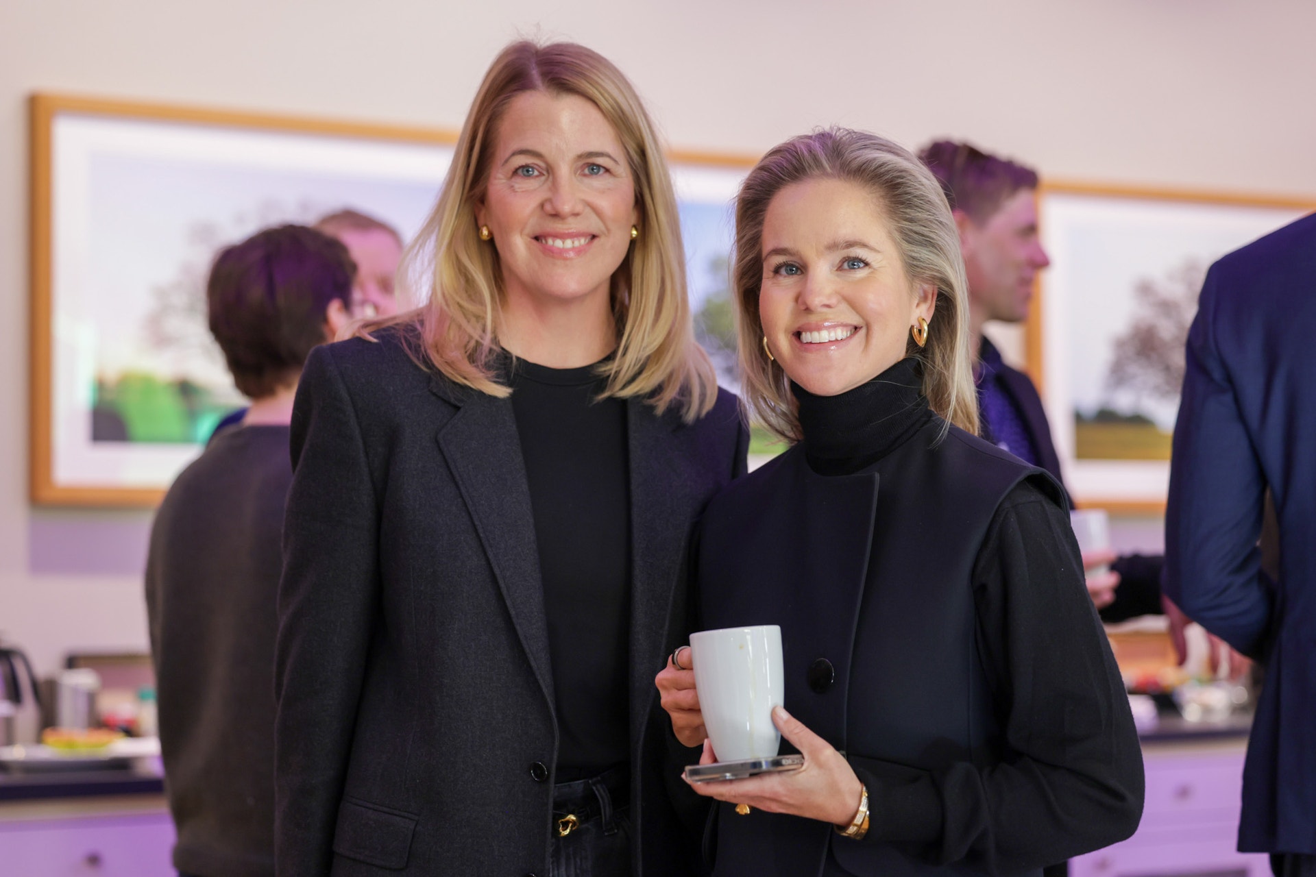 Två kvinnor i mörka blazers på ett inomhusevenemang. De står framför varandra och ler mot kameran.