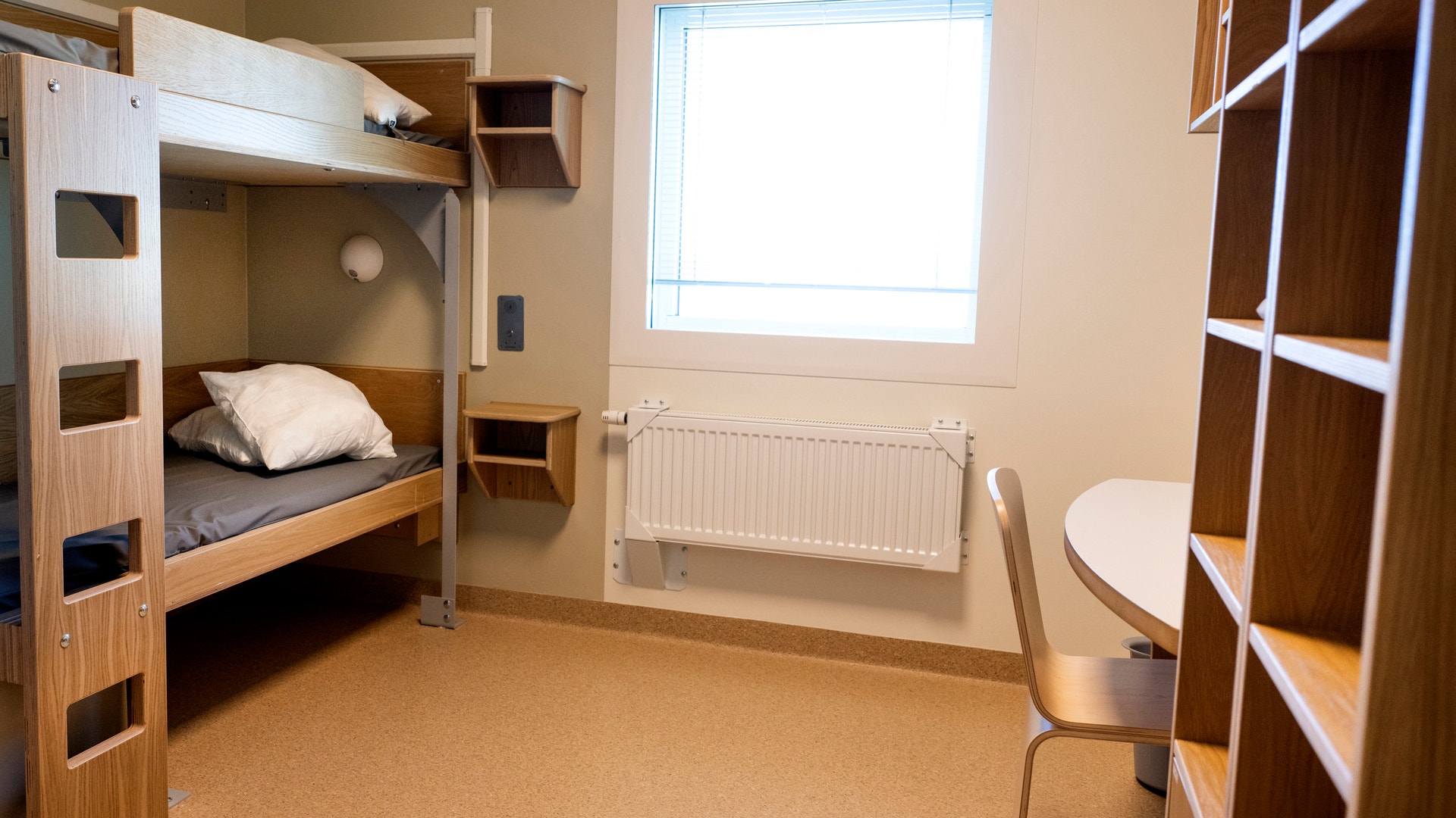 En enkel cell med våningssäng, bokhylla, skrivbord och stol i ljust trä. I väggen finns ett fönster.