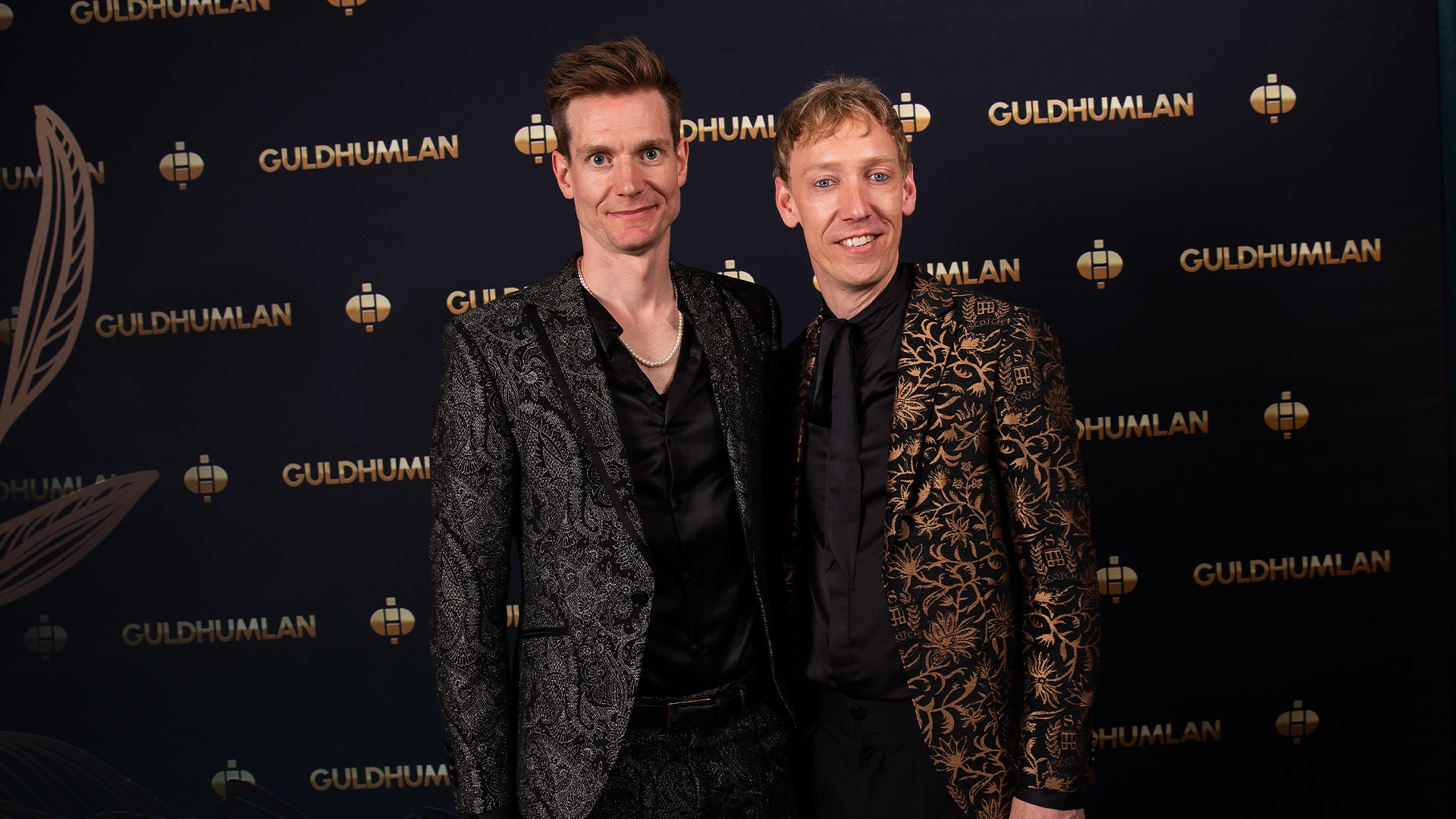 Två män poserar tillsammans framför en vägg med Guldhumlan-logotyper vid en event.