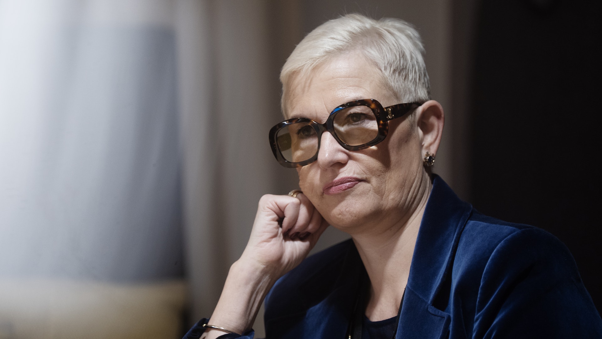 Korthårig blond kvinna med stora, mörka glasögon sitter med ansiktet lutat mot ena handen med ett allvarlig uttryck.