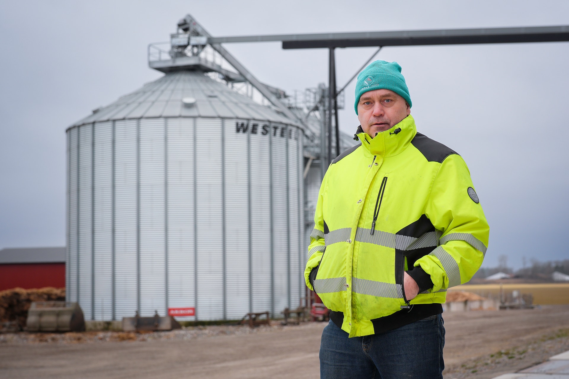 En person i gul säkerhetsjacka och mössa står framför en stor metallsilo på en jordbruksanläggning.