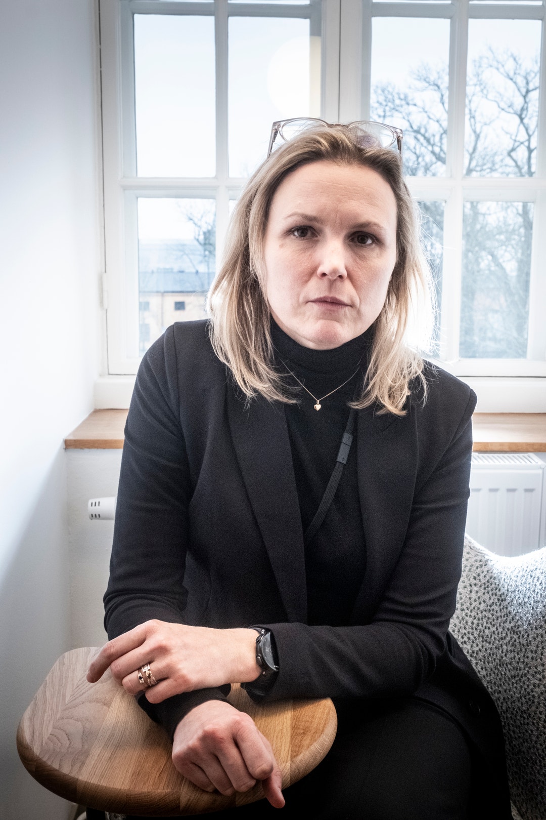 Porträtt av en kvinna i svarta kläder som sitter framför ett stort fönster med snöigt vinterlandskap i bakgrunden.