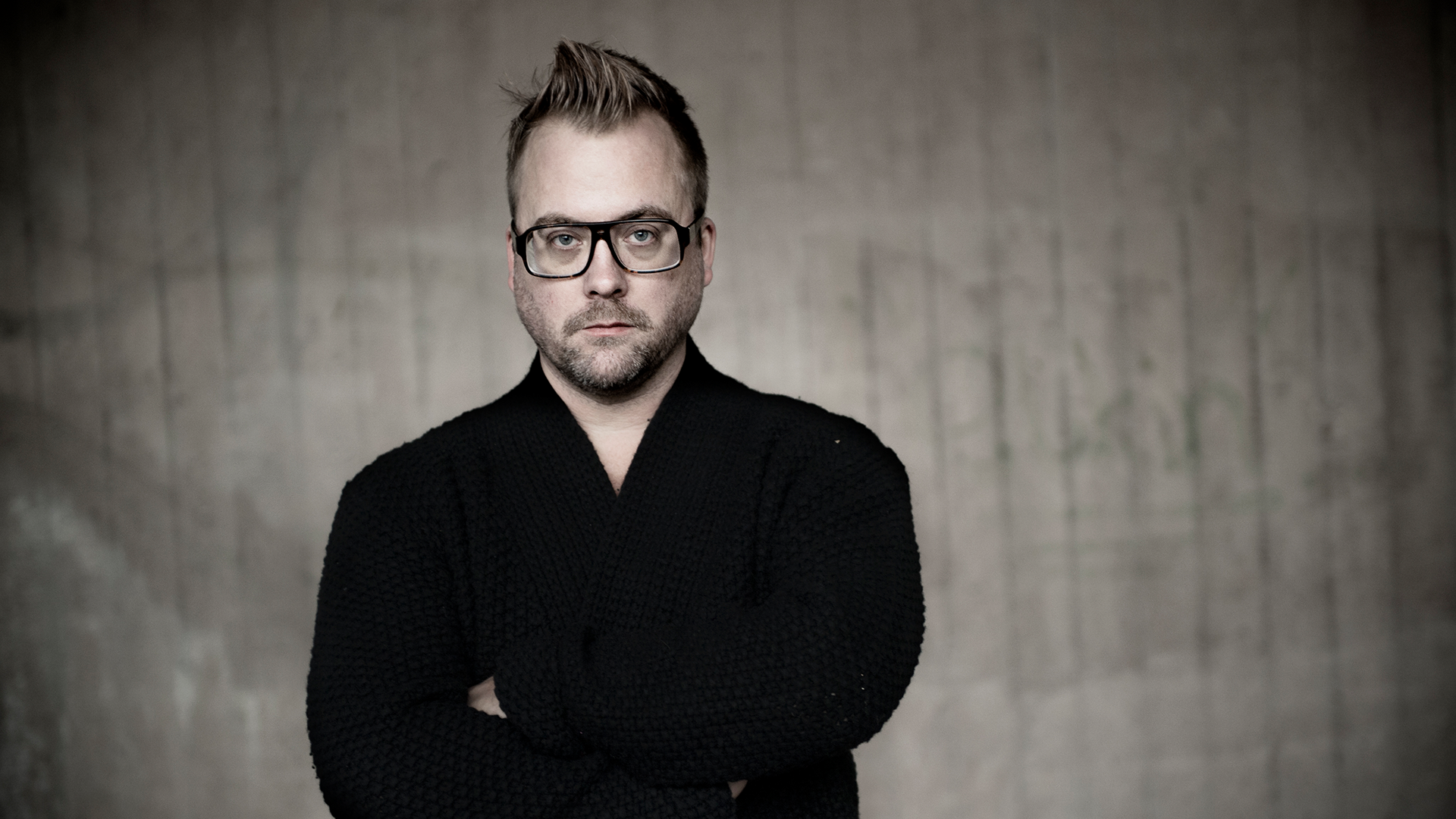 Mårten schultz står med armarna i kors och tittar allvarligt in i kameran. Han har en svart kofta och står poserande framför en betongvägg.