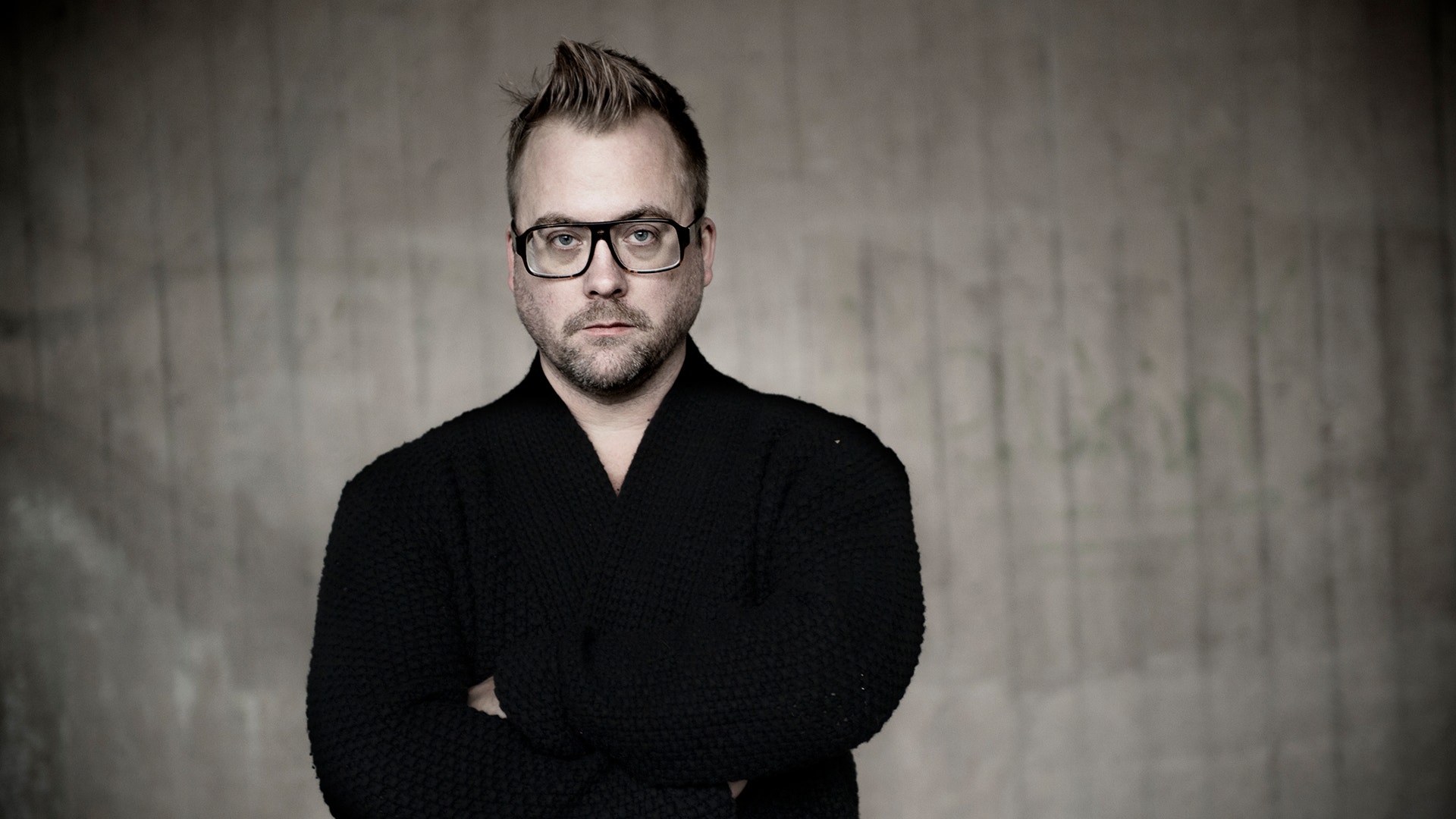Mårten schultz står med armarna i kors och tittar allvarligt in i kameran. Han har en svart kofta och står poserande framför en betongvägg.