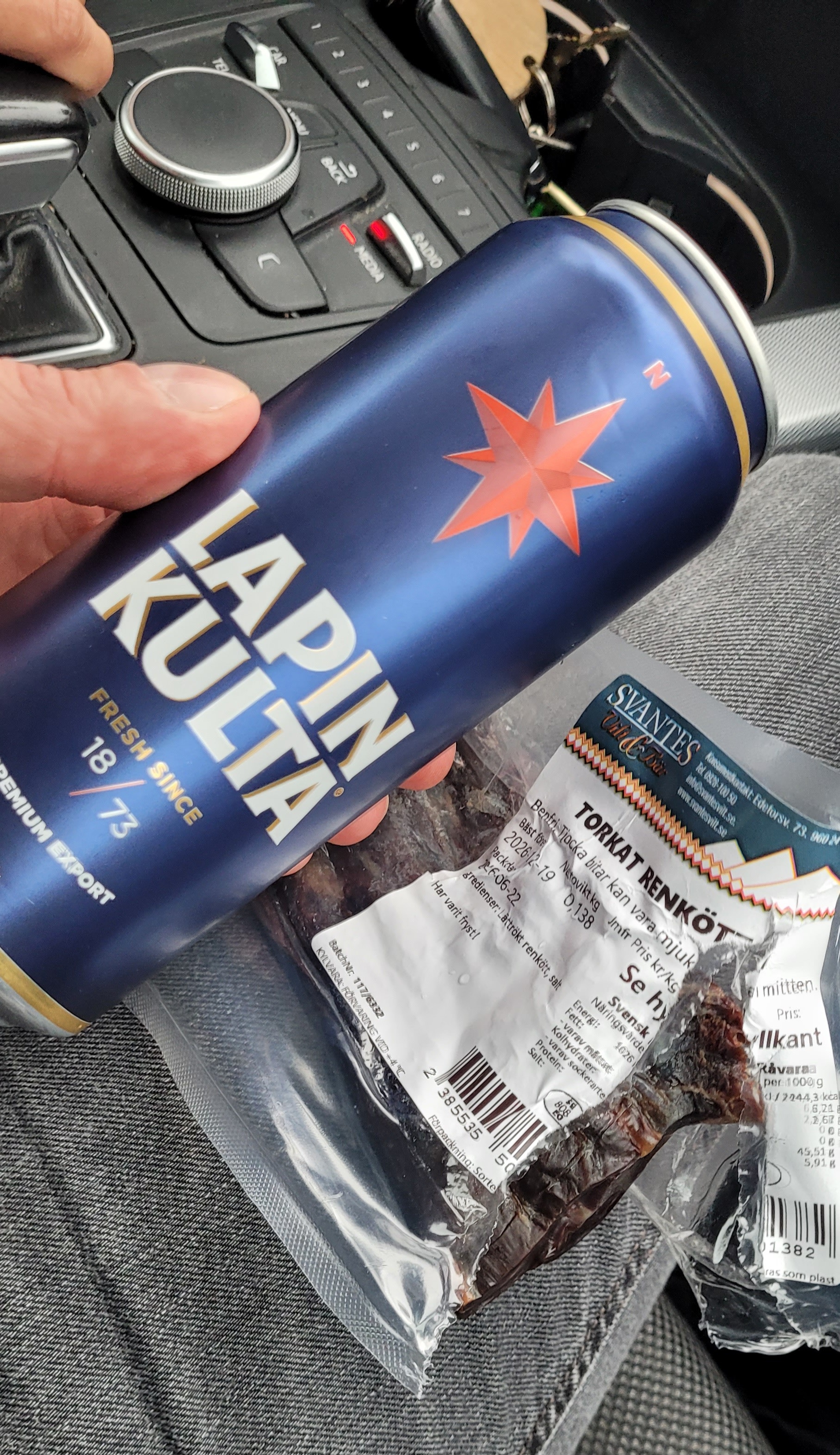 En hand håller en blå energidrycksburk med röd stjärna och texten Alpha inne i en bil. Bredvid burken ligger snackspaket.
