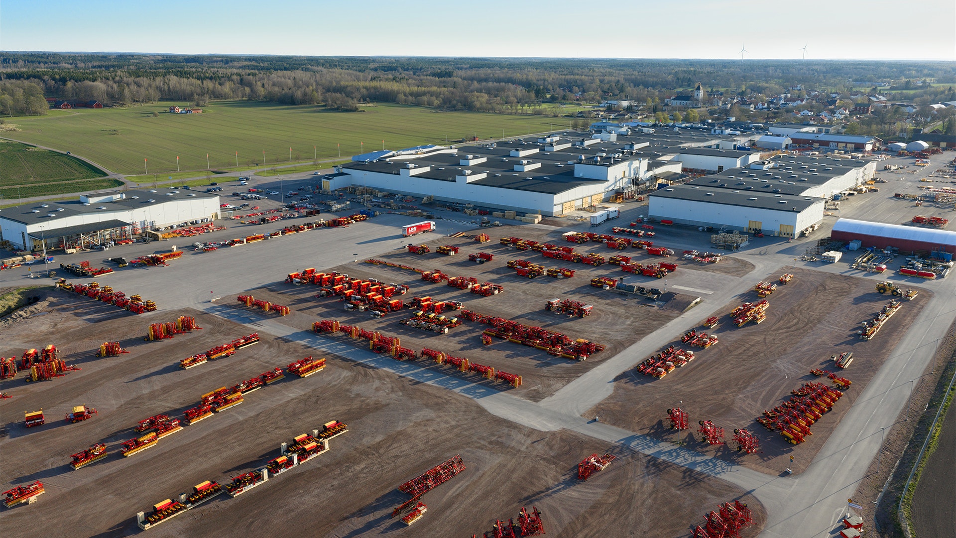 Flygfoto av ett industriområde med många röda jordbruksmaskiners parkerade i ordnade rader. I bakgrunden syns industribyggnader, gröna åkermarker och ett bostadsområde.