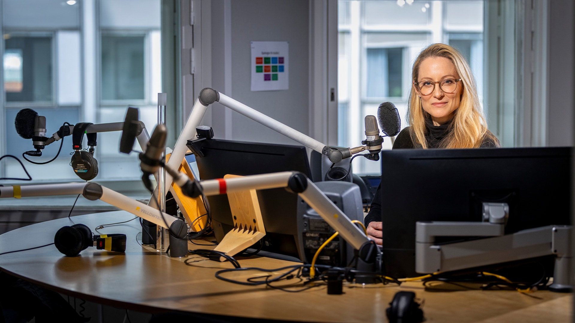 En kvinna sitter vid ett skrivbord i en modern inspelningsstudio omgiven av mikrofoner och annan teknisk utrustning för podcast eller radioinspelning.