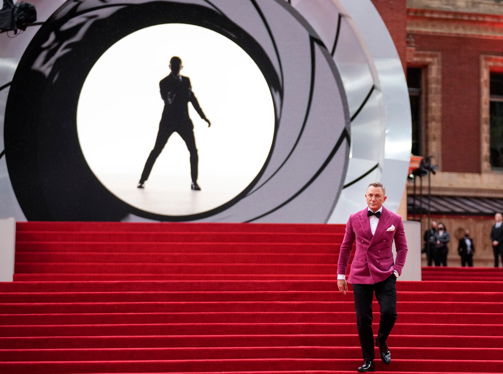 En man i rosa kavaj står på röda trappor framför en stor skärm som visar en agent i silhuett inom ett cirkulärt mål, en klassisk James Bond-scen.