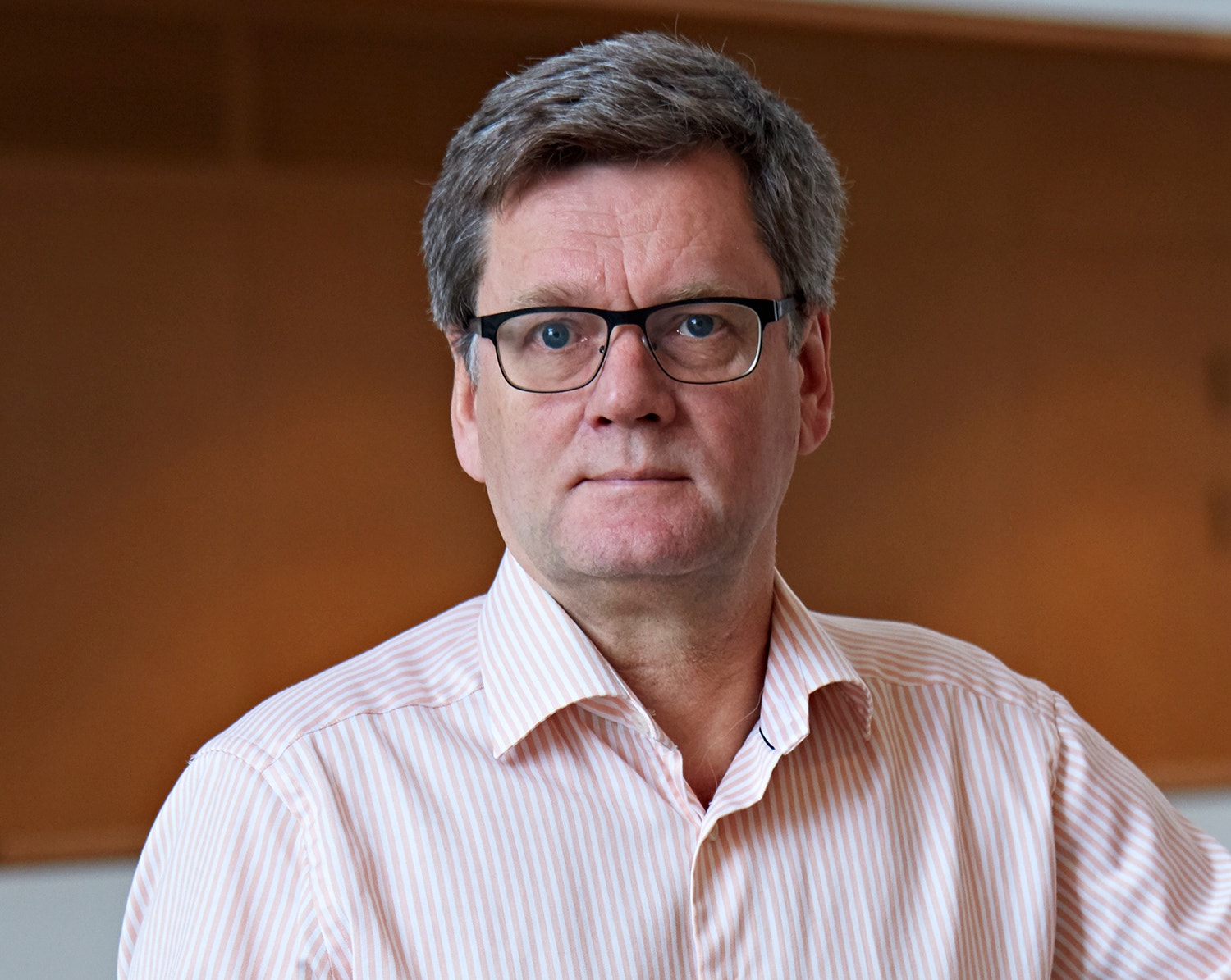 Porträttfoto av en man med glasögon och rosa randig skjorta mot en brun bakgrund.