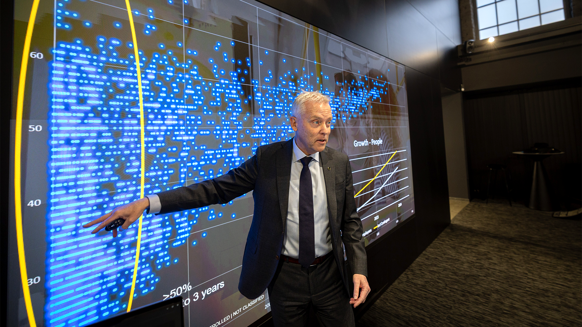Man i kostym presenterar framför en stor digital skärm med blå datavisualisering och grafer