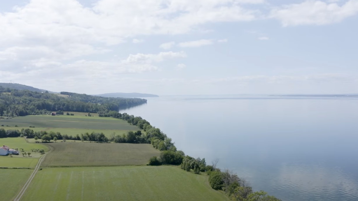 Flygfoto över Vättern med gröna jordbrukslandskap, skogar och lugnt vatten under molnig himmel.