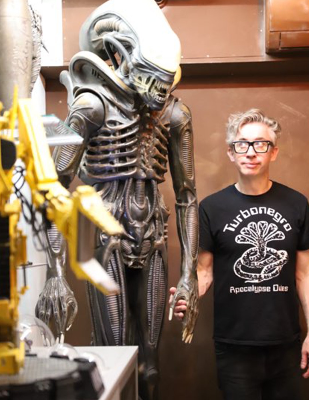 En person står bredvid en stor metallisk skulptur av en alien-varelse med långsträckt huvud och ribbestrukturer på kroppen.