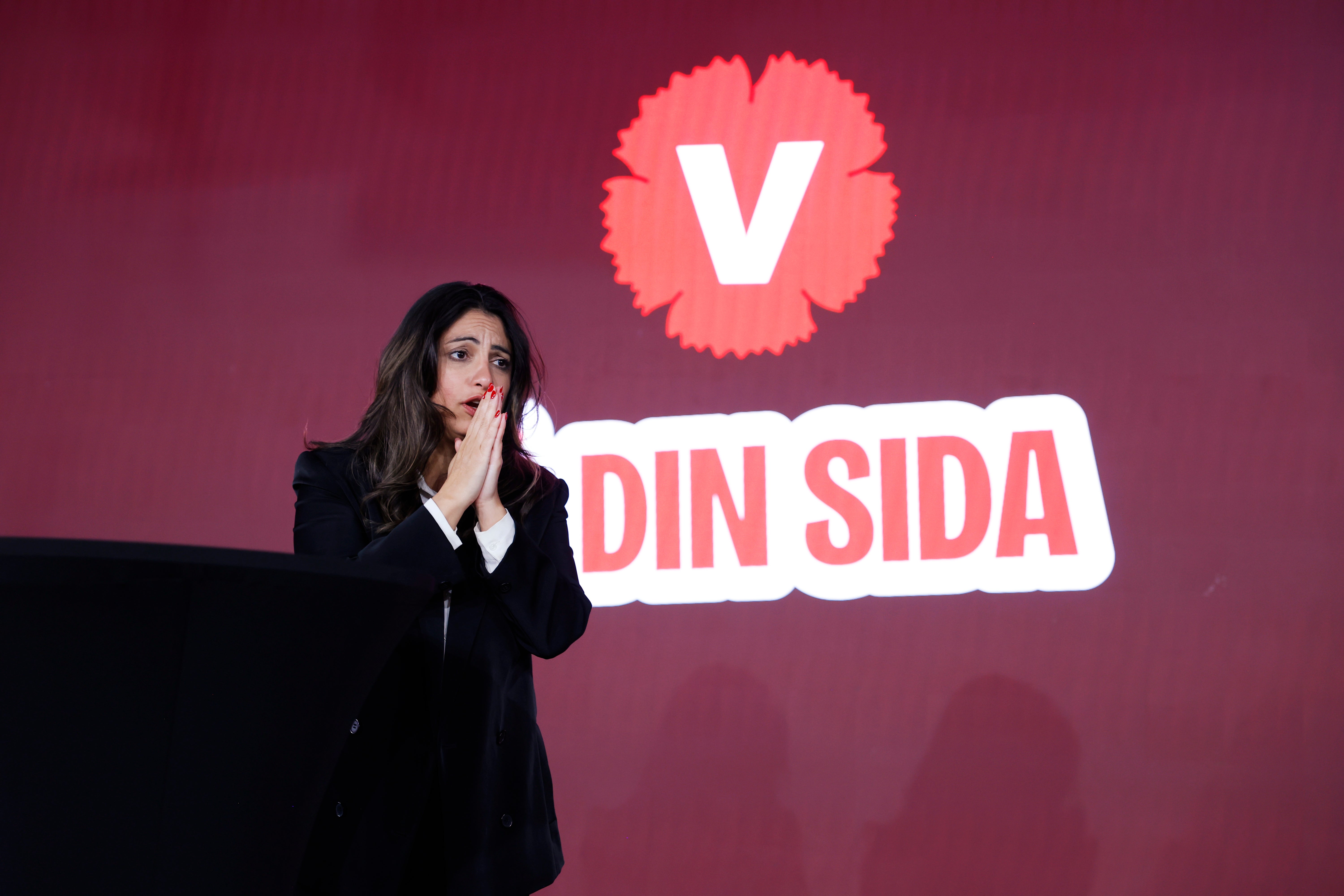 En kvinna står vid en talarstol på en scen med rörda händer framför ansiktet. Bakom henne syns Vänsterpartiets logotyp och texten 'Din sida'.