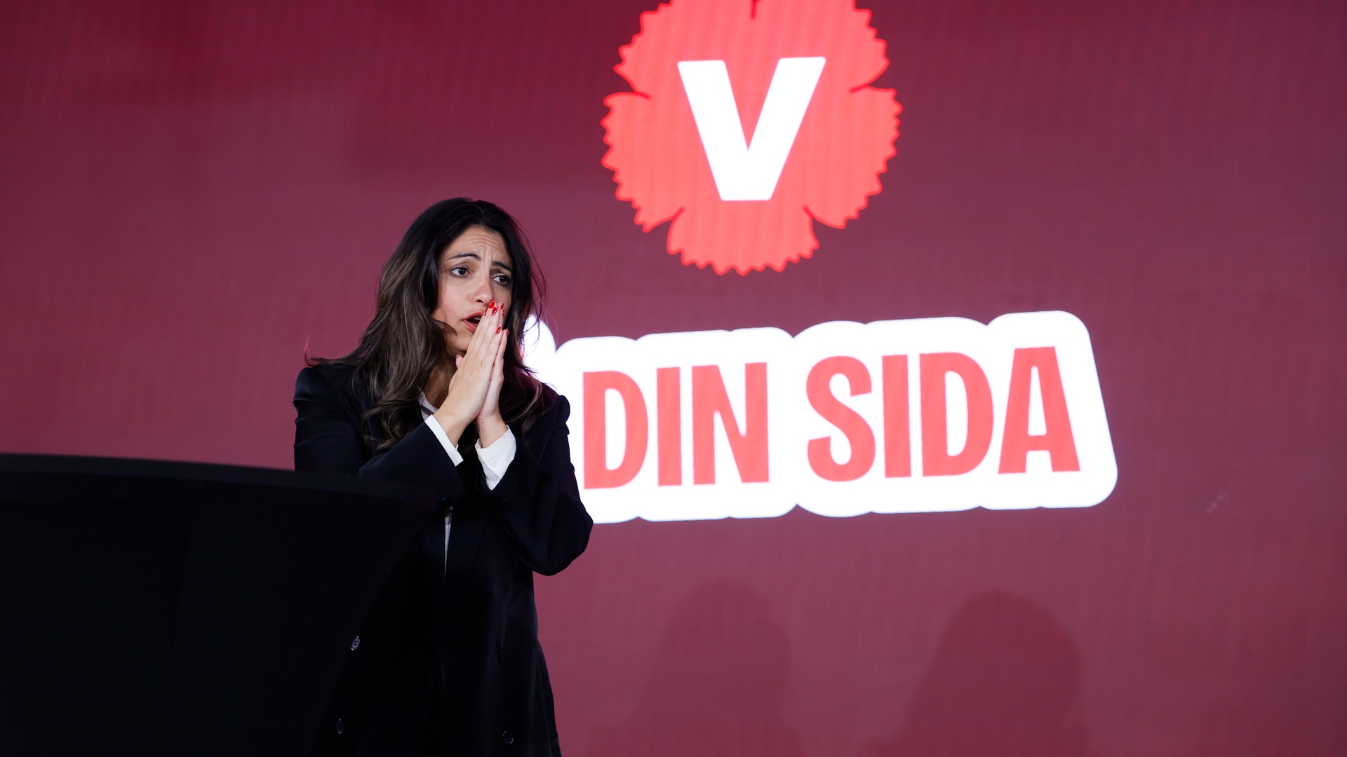 En kvinna står vid en talarstol på en scen med rörda händer framför ansiktet. Bakom henne syns Vänsterpartiets logotyp och texten 'Din sida'.
