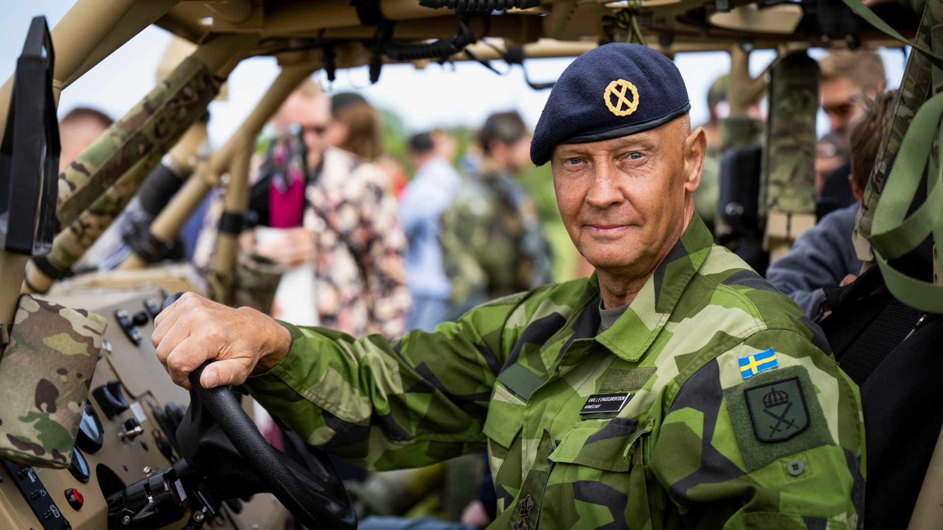 En militär officer i grön kamouflageuniform och blå baskermössa står vid ett militärfordon under en utställning.