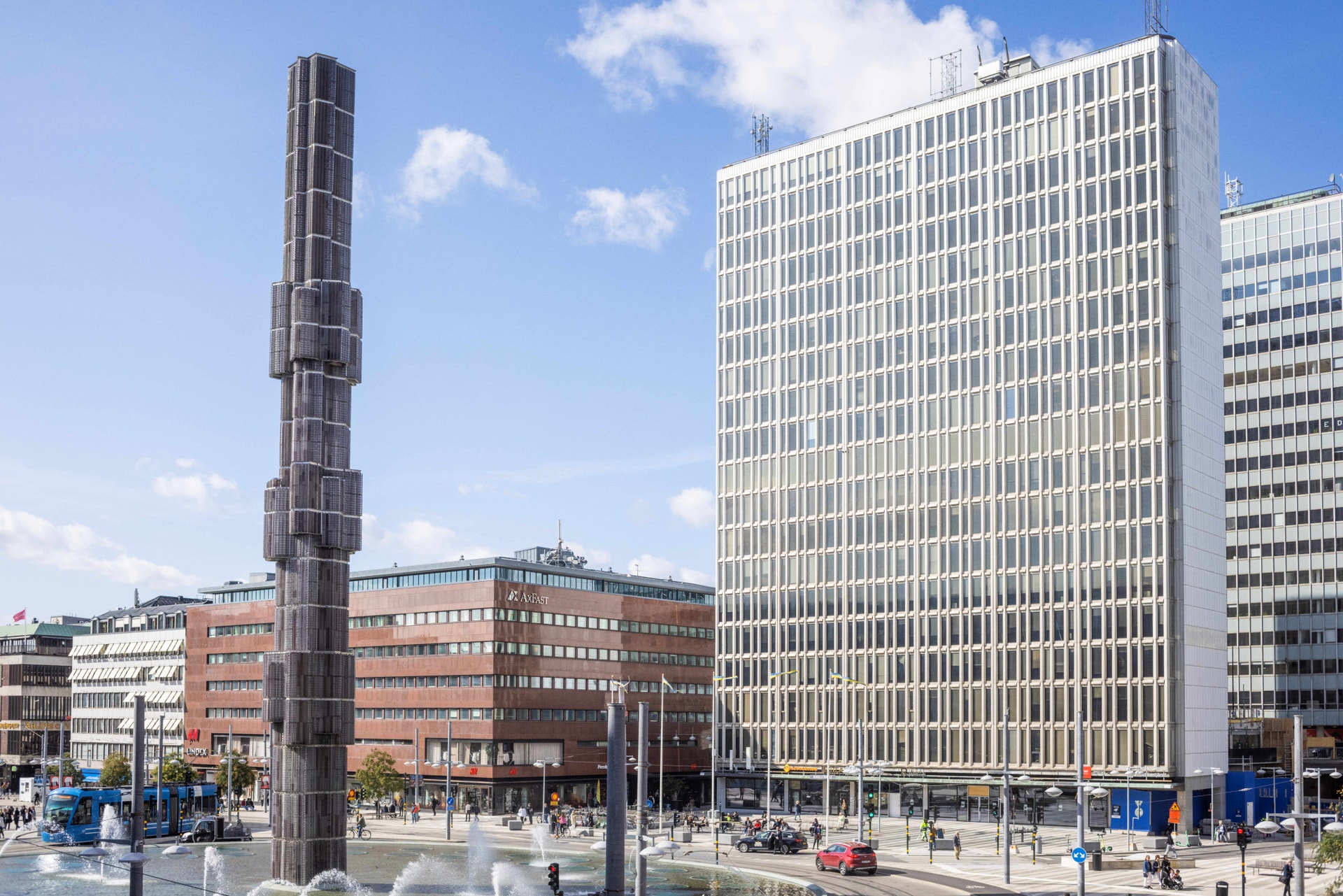 Sergels torg i Stockholm med en hög cylindrisk monument i förgrunden och moderna kontorsbyggnader i bakgrunden under blå himmel.