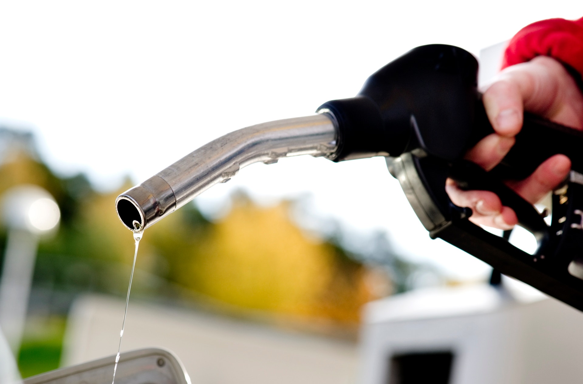En hand håller en bensinpump med röd och svart handtag. Bränsle droppar från munstycket. I bakgrunden syns en suddig tankstation.