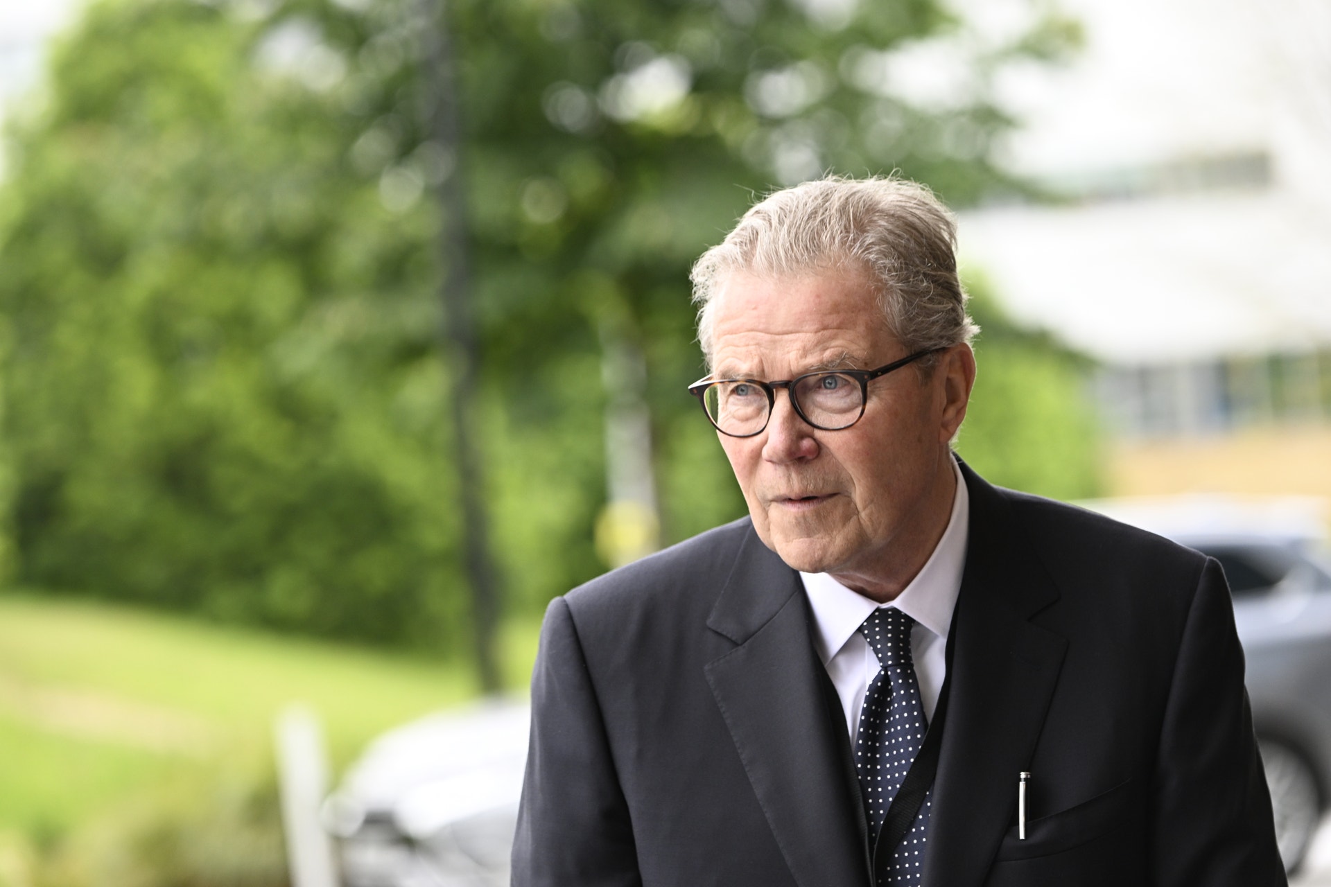Porträtt av en äldre man i kostym och glasögon utomhus med gröna träd i bakgrunden.