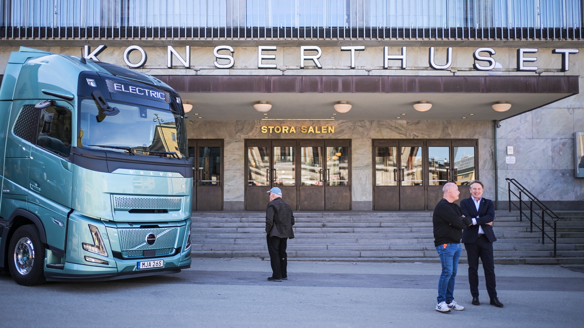 En blå Volvo elektrisk lastbil parkerad framför Konserthuset. Några personer står på platsen framför byggnadens entré.