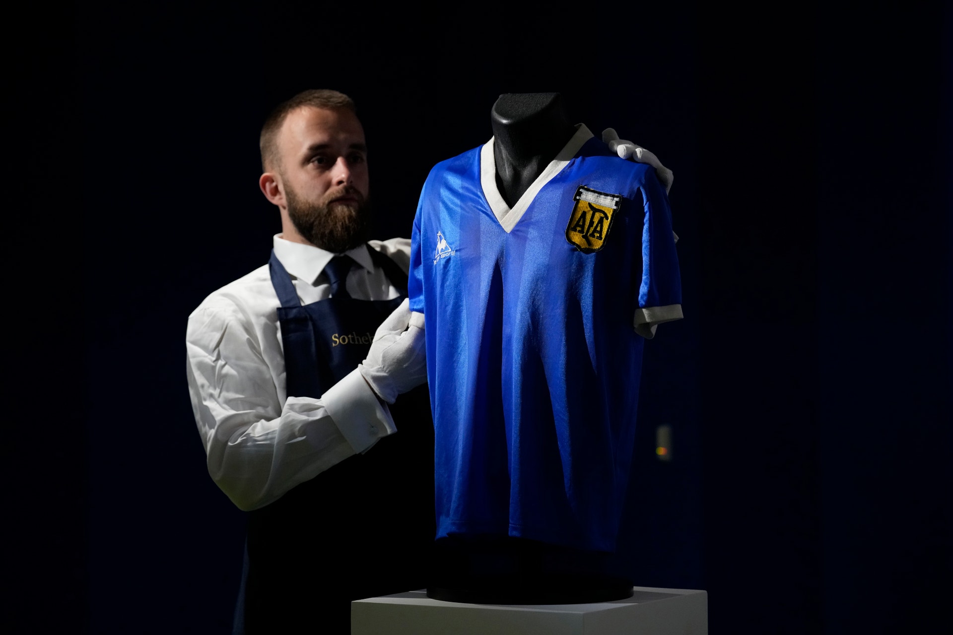 En man presenterar en blå fotbollströja på en skyltdocka. Han bär vita handskar och vit skjorta med svart väst.