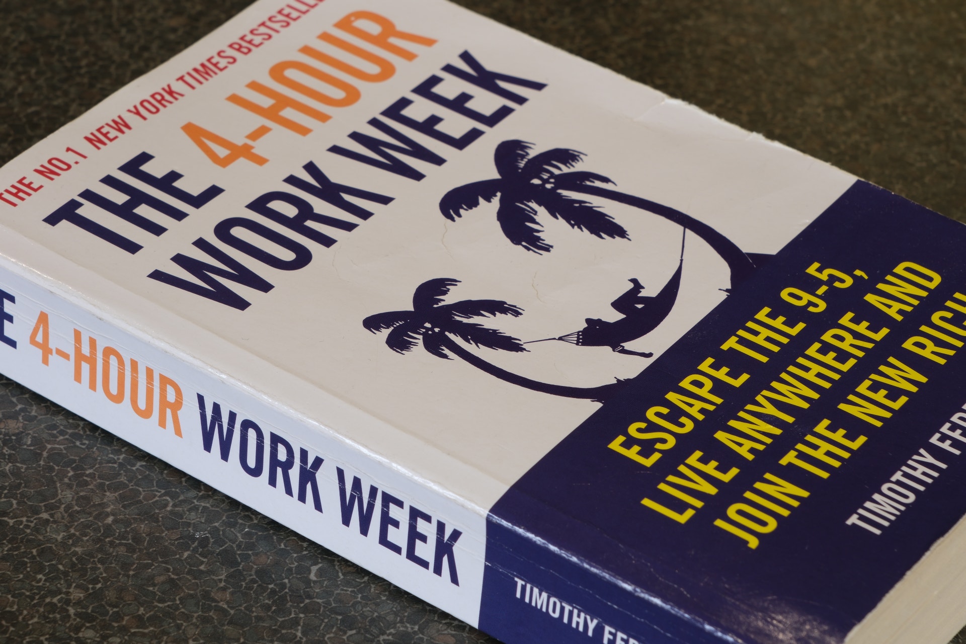 Boken The 4-Hour Workweek av Timothy Ferriss ligger på ett mörkt underlag. Omslaget visar palmträd och texten är i orange och mörkblå färger.