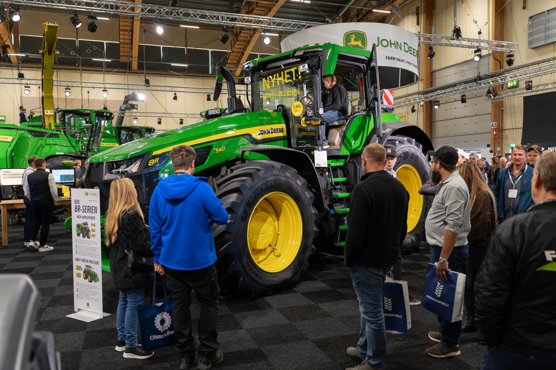 Besökare på en lantbruksmässa tittar på en grön John Deere-traktor som står utställd i en stor hall.