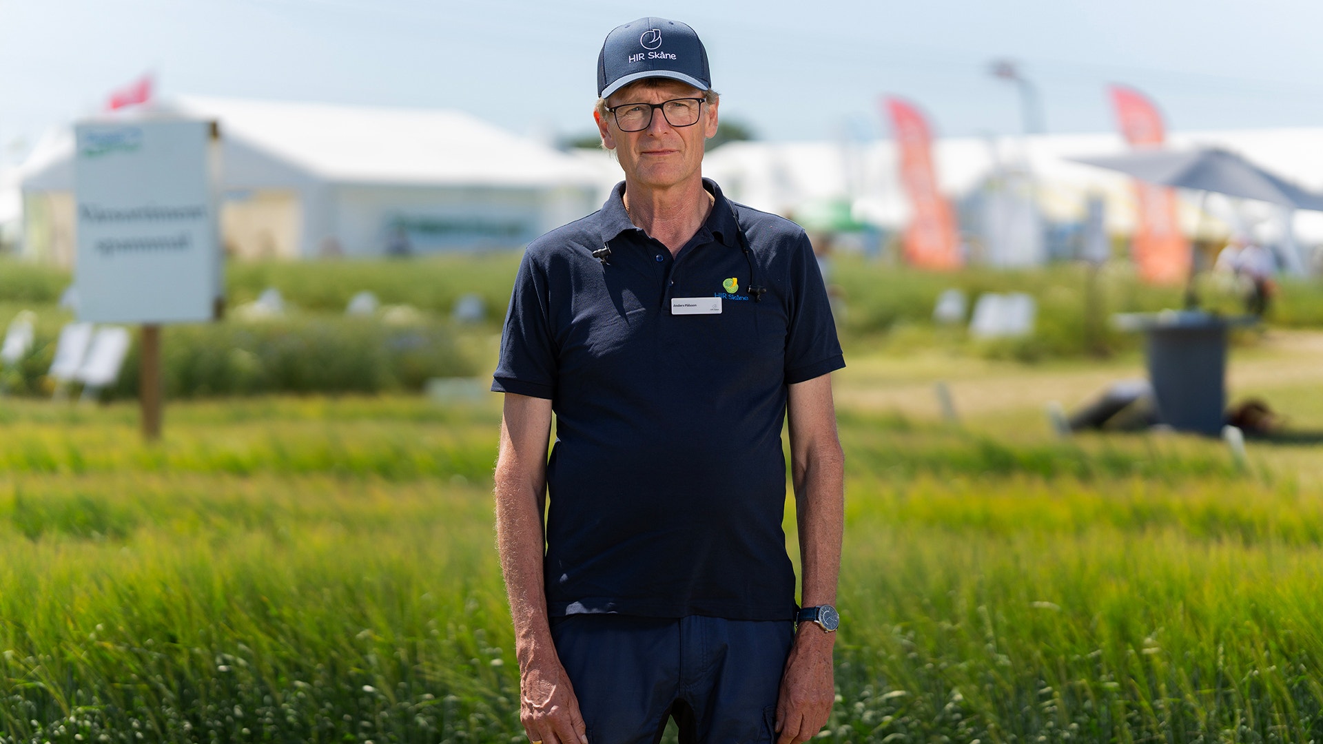Man i mörkblå arbetsuniform och keps står i ett grönt fält med industribyggnader i bakgrunden.
