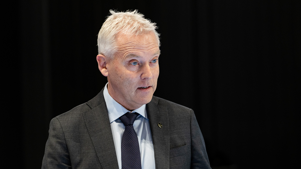 Porträtt av en äldre man i mörkgrå kostym och mörkblå slips mot svart bakgrund.