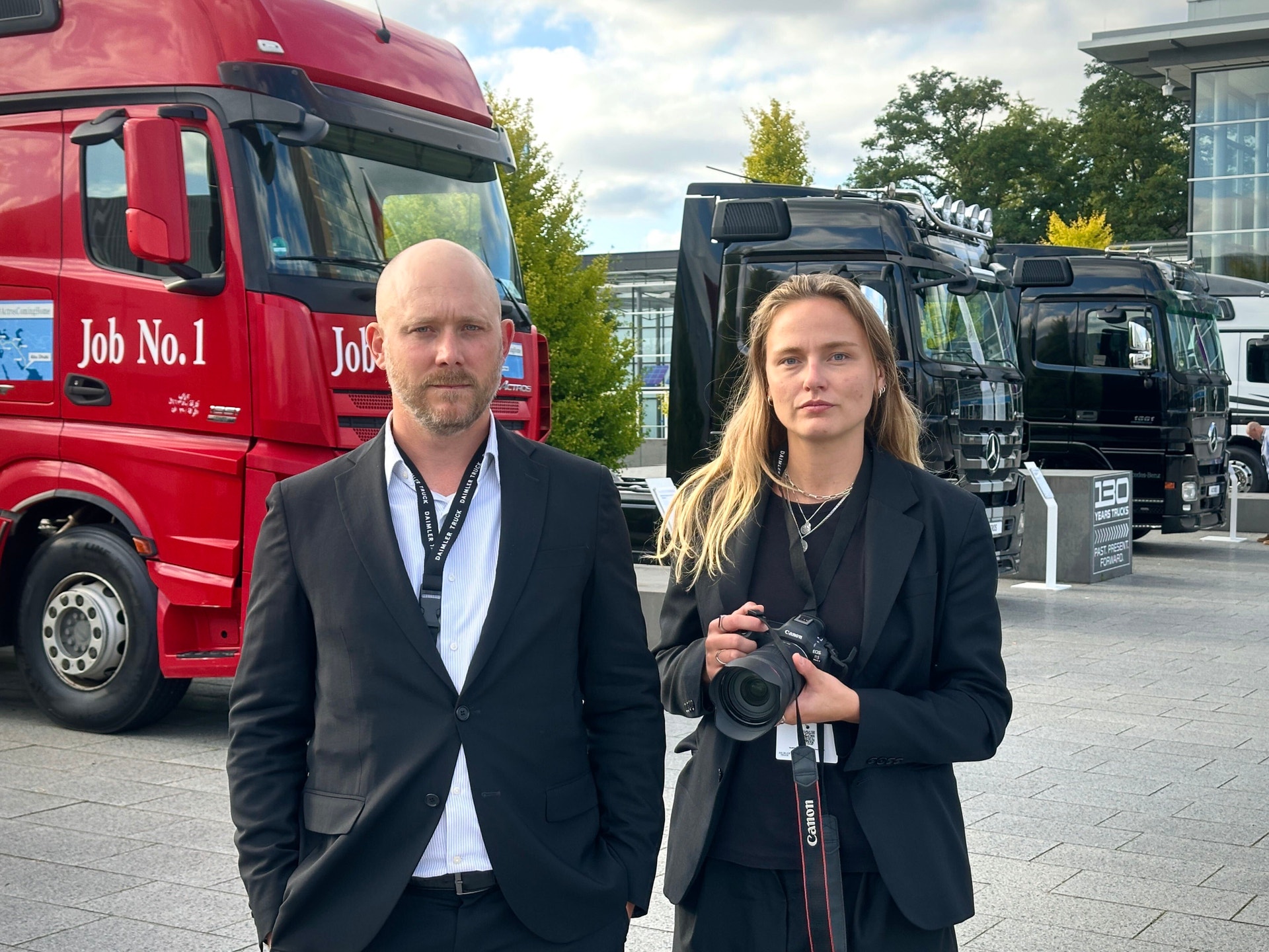 En man och en kvinna, klädda i kostym, står framför flera lastbilar utomhus. Kvinnan håller en kamera.