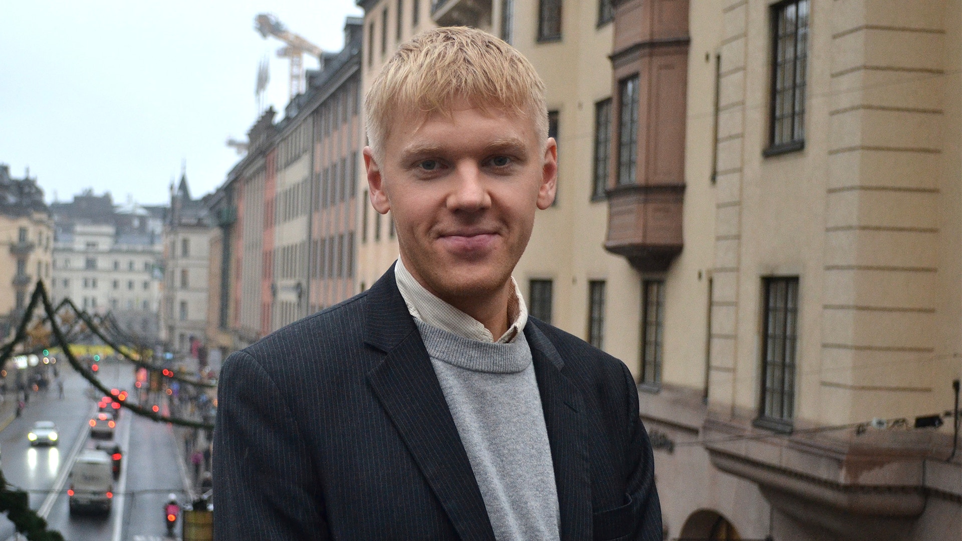 Porträtt av en man med blont hår, klädd i mörk kavaj och grå polotröja, fotograferad framför klassiska europeiska byggnader.