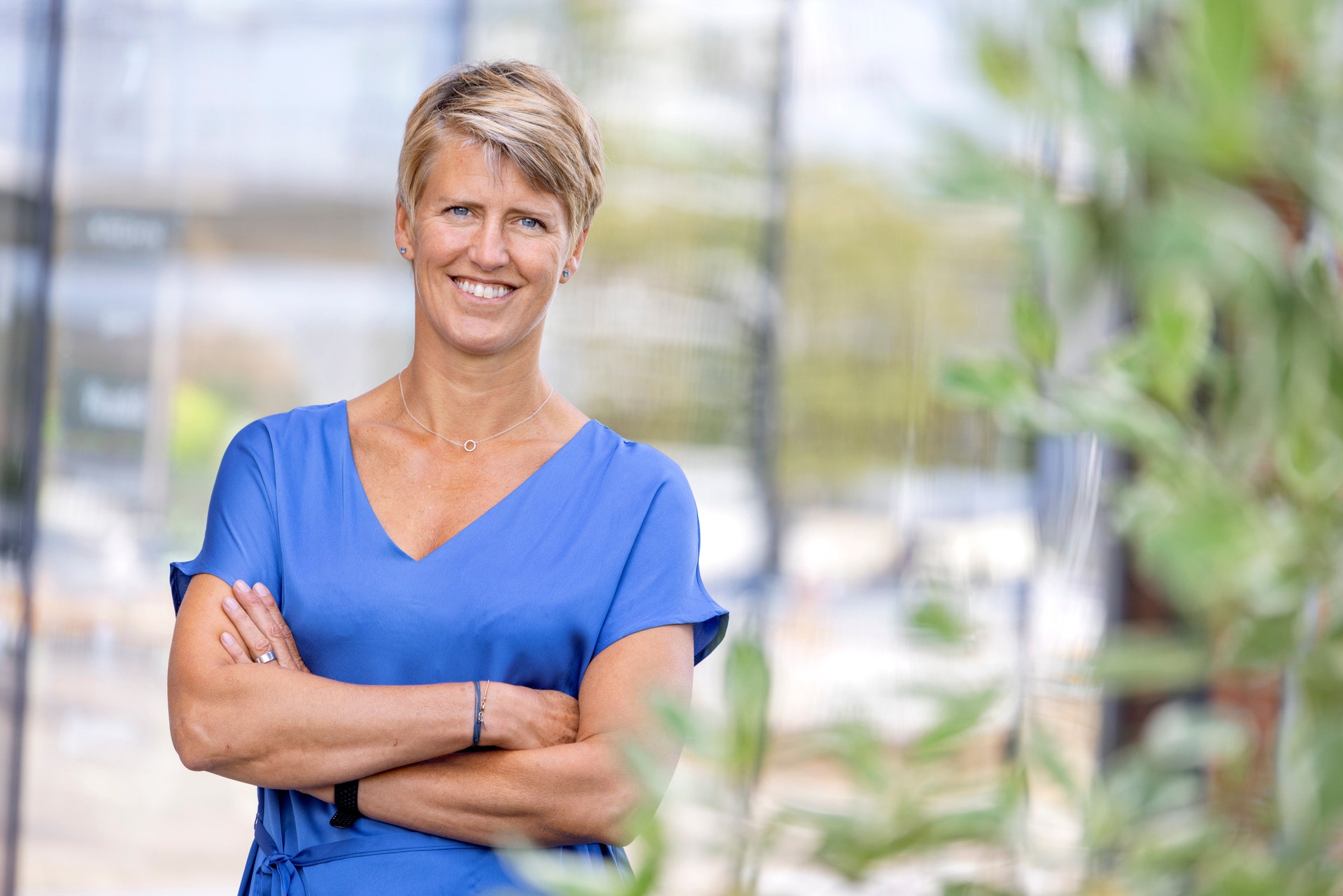 Leende kvinna med kort blont hår och blå klänning står med korsade armar utomhus.