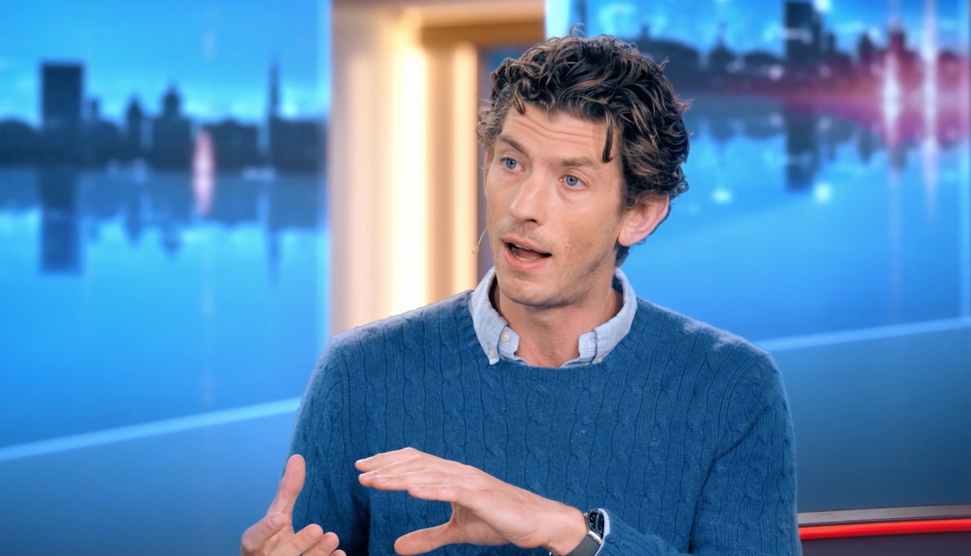 En man i blå tröja pratar och gestikulerar i en tv-studio.