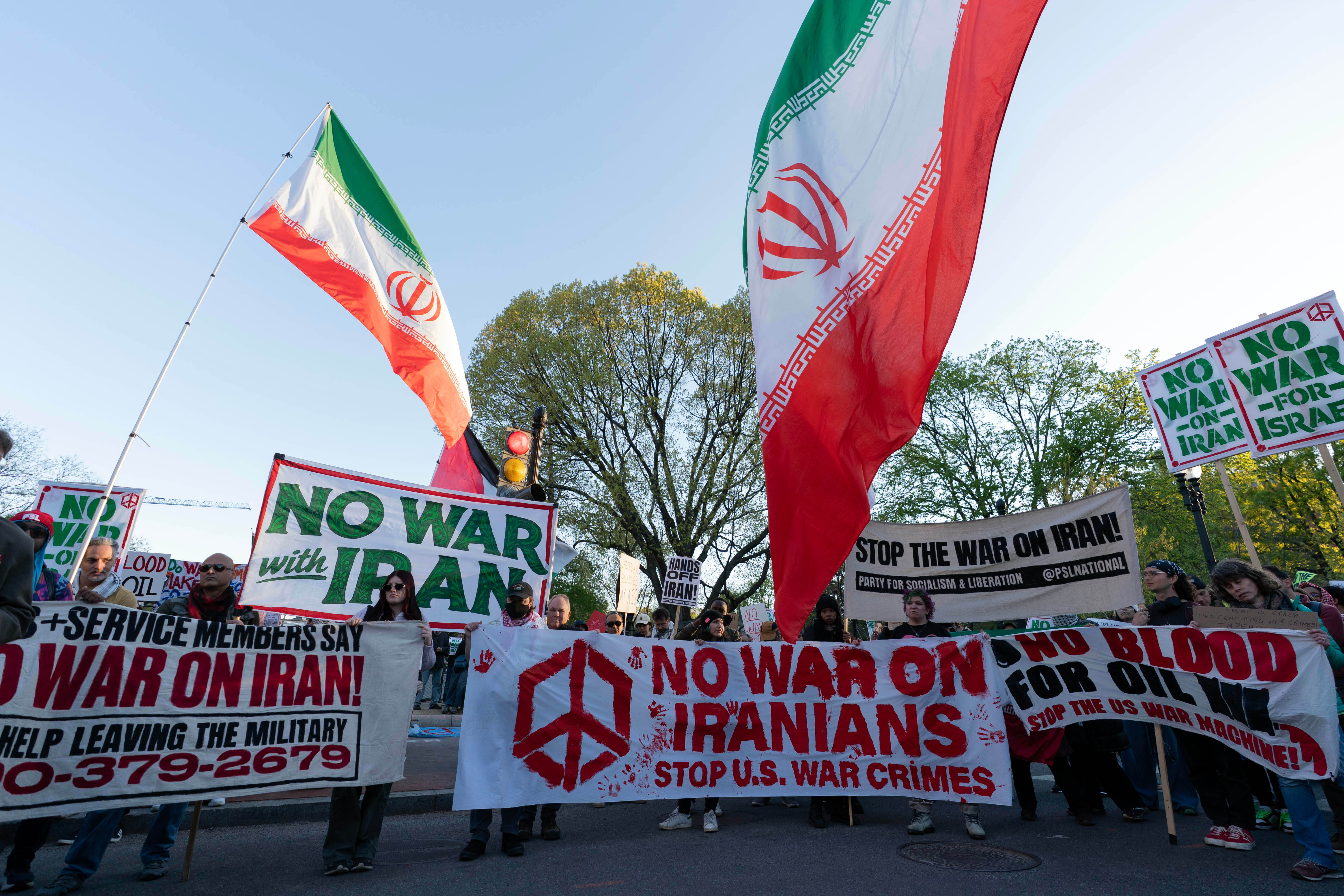 Fredsdemonstranter håller iranska flaggor och plakat mot krig. Banderoller visar budskap som "No War on Iran" och "Stop U.S. War Crimes".