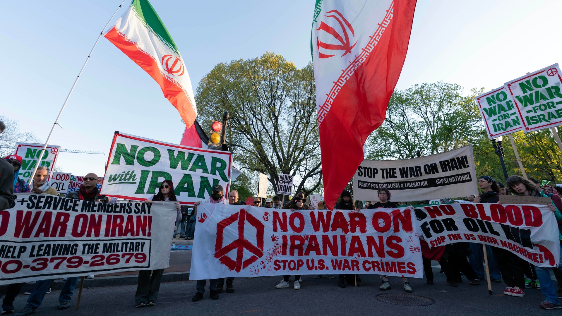 Fredsdemonstranter håller iranska flaggor och plakat mot krig. Banderoller visar budskap som "No War on Iran" och "Stop U.S. War Crimes".