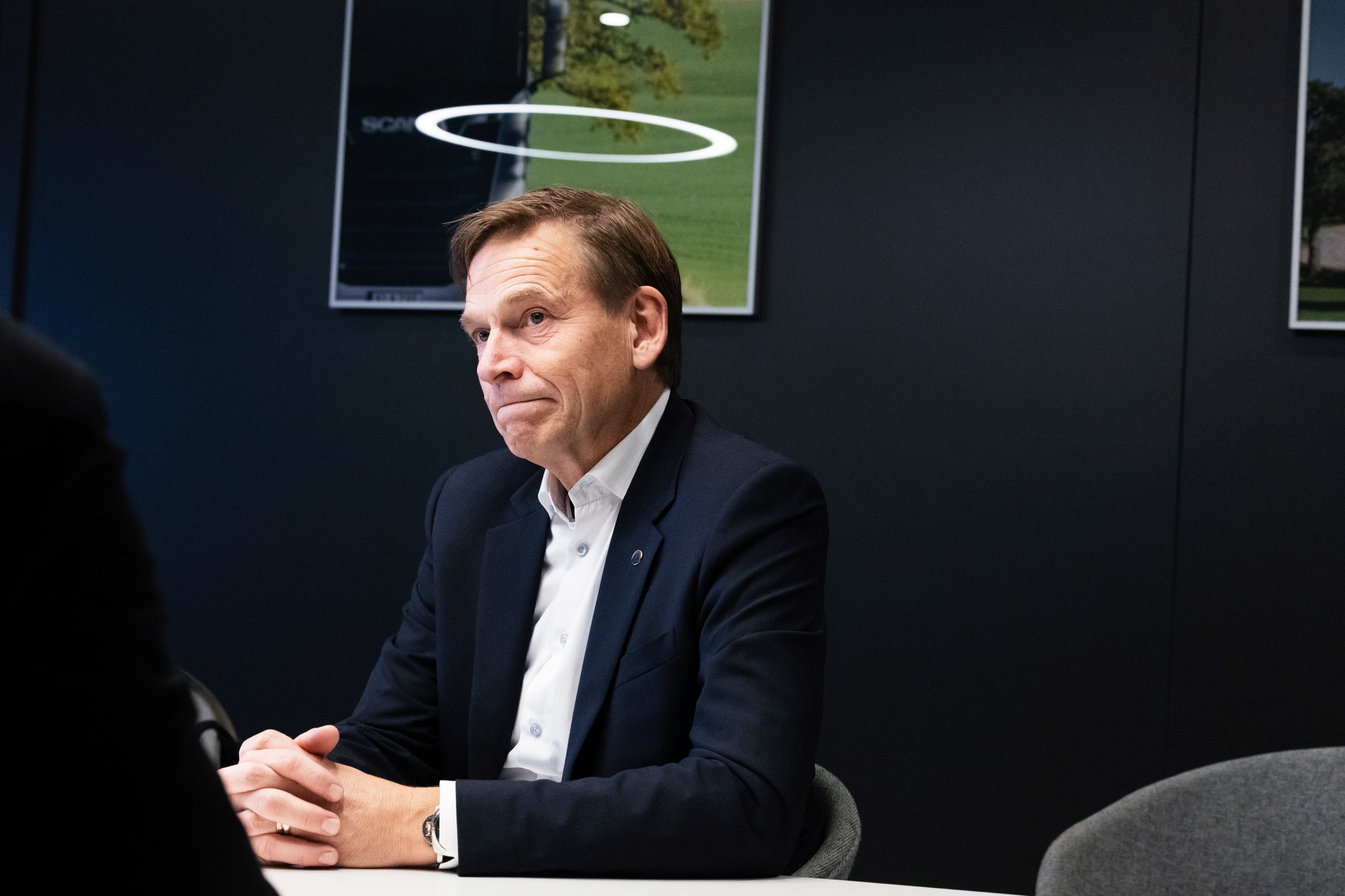 En man i kostym sitter vid ett bord under en intervju, med en tavla i bakgrunden.